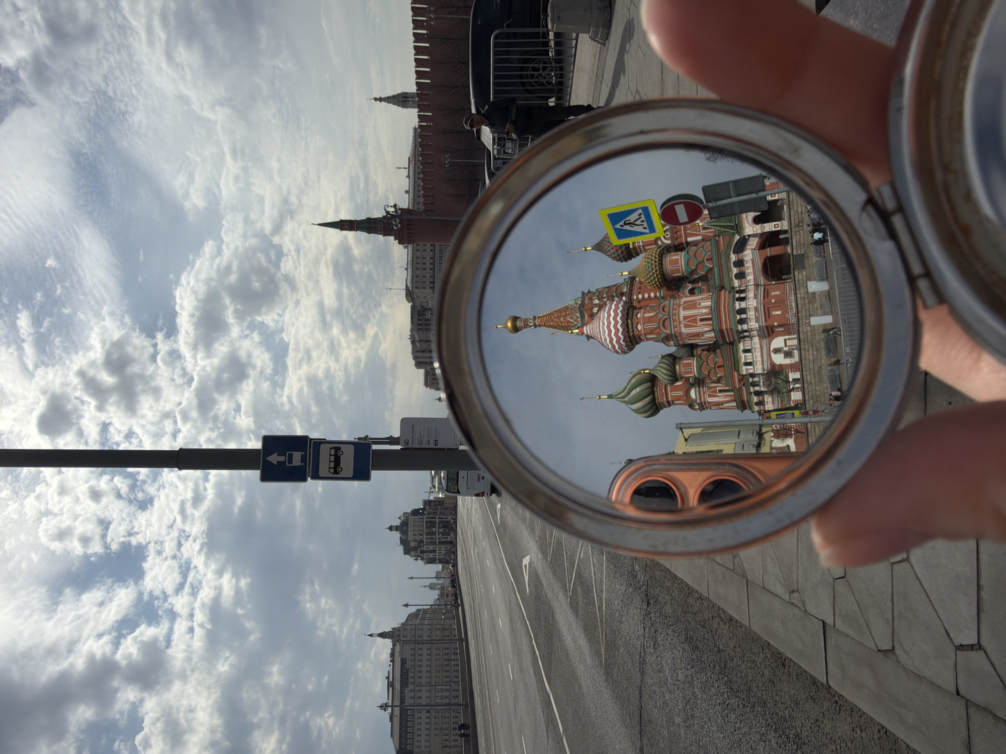 The image depicts a view of St. Basil's Cathedral in Moscow, Russia, as reflected in a small round mirror held in someone's hand. The cathedral is prominently featured in the mirror's reflection, showcasing its colorful domes and intricate architectural details. Outside the mirror's reflection, parts of the Moscow Kremlin wall and towers can be seen, along with a cloudy sky above. The foreground includes elements of the city street, such as a bus stop sign and pedestrian crosswalk sign. The photograph seems to be taken at ground level, showing a portion of the paved walkway beside the street.