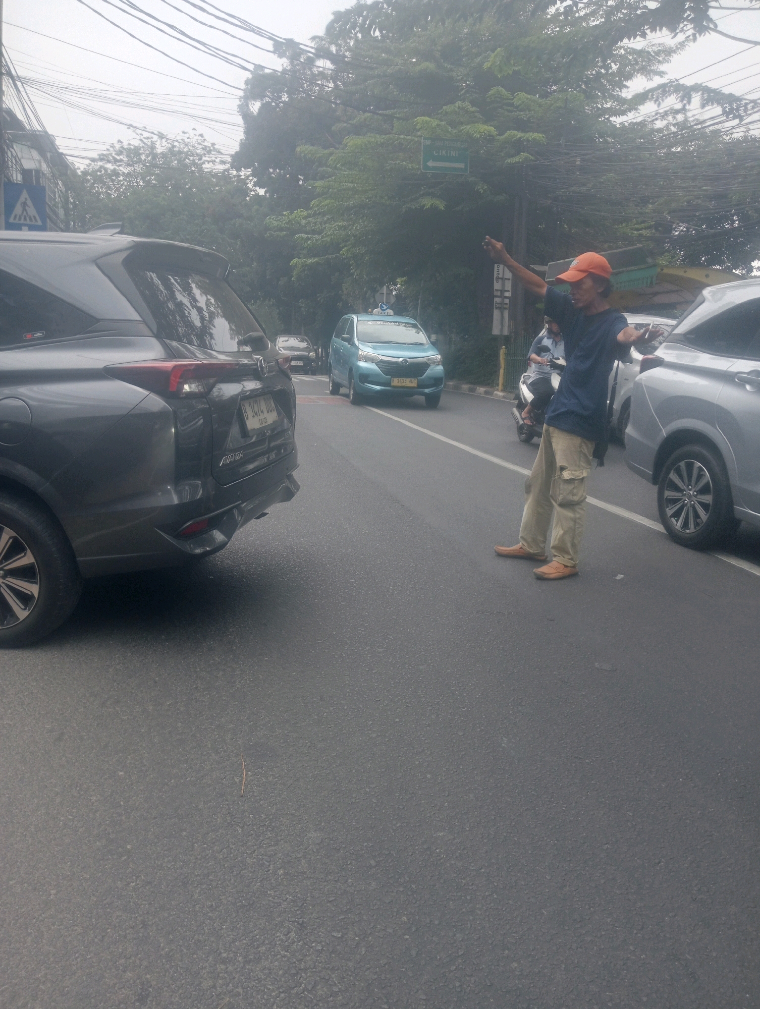 In South Jakarta, Indonesia, on a foggy day, a man wearing an orange cap and a dark blue shirt stands on the right side of the road, pointing forward with his right hand. He is wearing light brown cargo pants and brown shoes. In front of him, a silver car is parked, and a blue car is driving past. To the left, a dark gray SUV is also on the road. Overhead, numerous electrical wires are visible, and green trees line the background. A street sign with green lettering is partially obscured by the trees. The road appears to be wet, suggesting recent rain or high humidity.