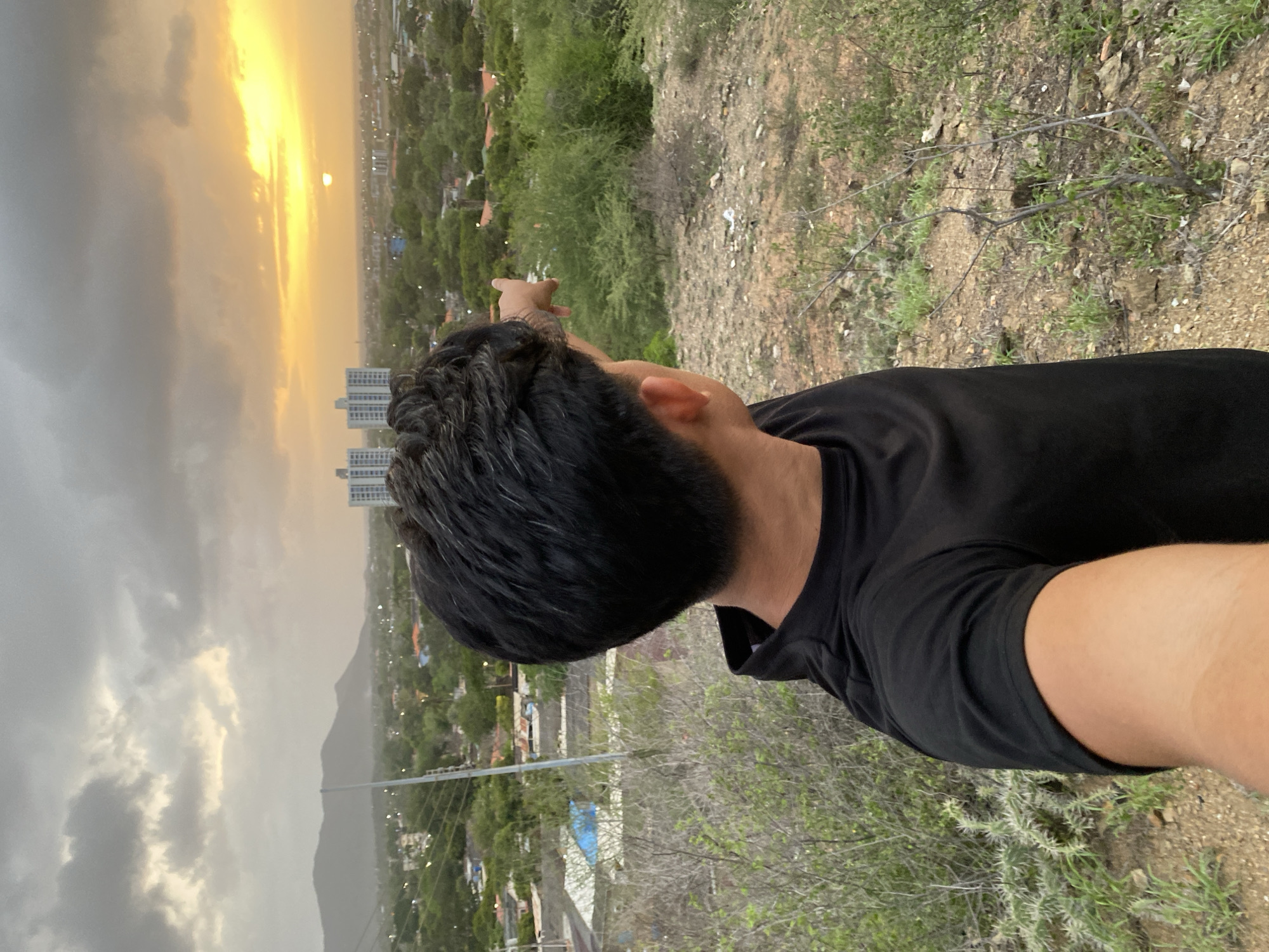A young man with dark hair, wearing a black t-shirt, stands on a hill overlooking the city of Juan Griego, Venezuela. He is facing away from the camera, looking towards the horizon where the sun is setting. His right arm is extended, and he is pointing towards the distant landscape. The sky is filled with dramatic clouds, illuminated by the warm glow of the setting sun, which appears as a small bright orb near the horizon. Below, the city of Juan Griego is spread out, with buildings, trees, and roads visible. The foreground shows a rugged, dry terrain with sparse vegetation. The overall scene captures a moment of contemplation and appreciation of the natural beauty of the Venezuelan coastline at dusk.
