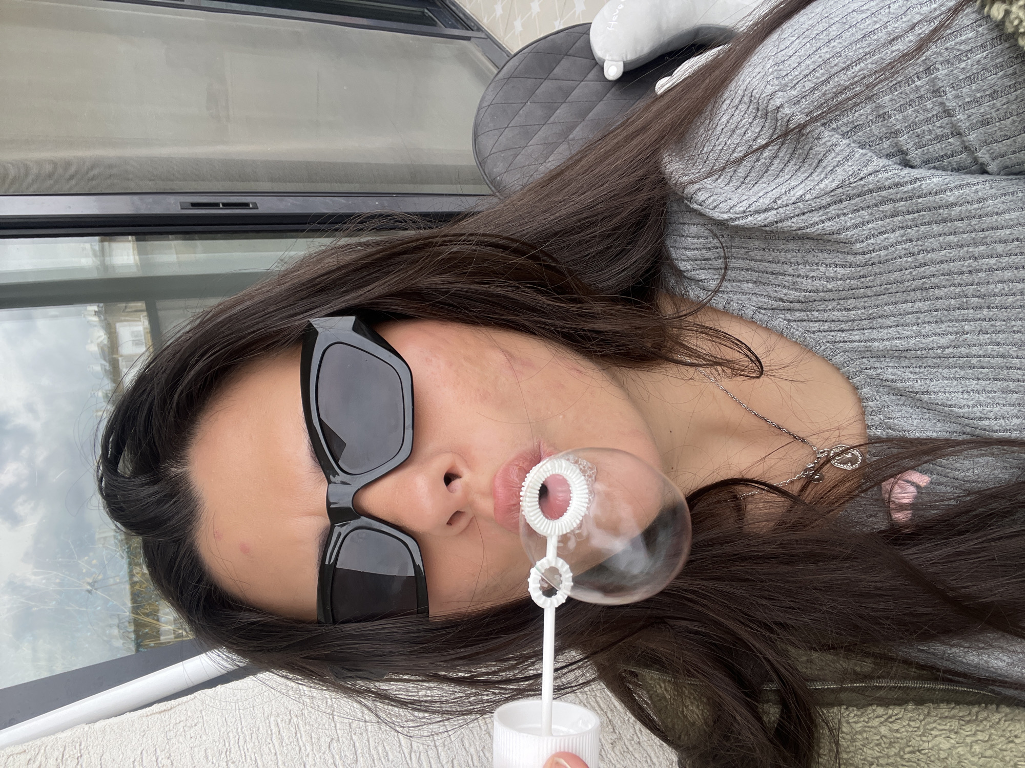 The image presents a close-up, chest-up view of a female individual on what appears to be an outdoor balcony in Kiryat Bialik, Israel. The subject, with long, dark brown hair, is wearing dark, angular sunglasses. Her attire includes a light grey, ribbed long-sleeve top and a textured olive-green jacket. A silver necklace with multiple pendants is visible around her neck. She is actively blowing a large, spherical soap bubble from a white plastic bubble wand, which has two distinct circular rings at its blowing end, one of which is currently forming the bubble. Minor skin blemishes are visible on her forehead and cheeks. The background on the left consists of a white, textured wall and a narrow white vertical element. To the right, a dark-framed glass sliding door or window reflects portions of residential buildings and a cloudy sky. A dark grey quilted chair and a segment of a light-colored, patterned floor are visible in the lower right quadrant.