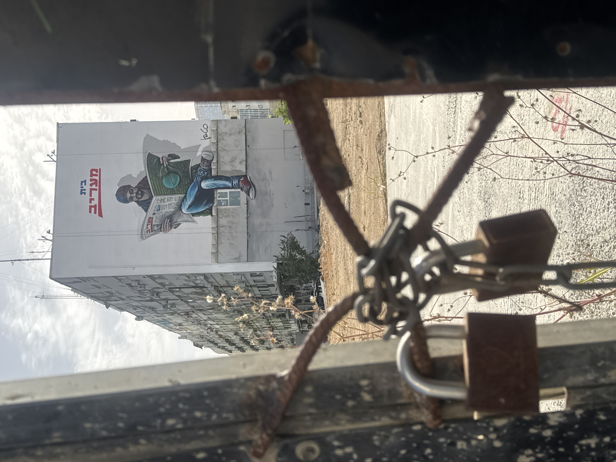 The image shows a locked metal gate, with multiple locks and chains, in the foreground. Through the gate, a building is visible with a large mural of a man reading a newspaper. The mural includes the text "Maariv House." The environment appears to be a vacant lot or construction area adjacent to the building. Based on the reference to "Maariv House," the location is likely in Tel Aviv, Israel.
