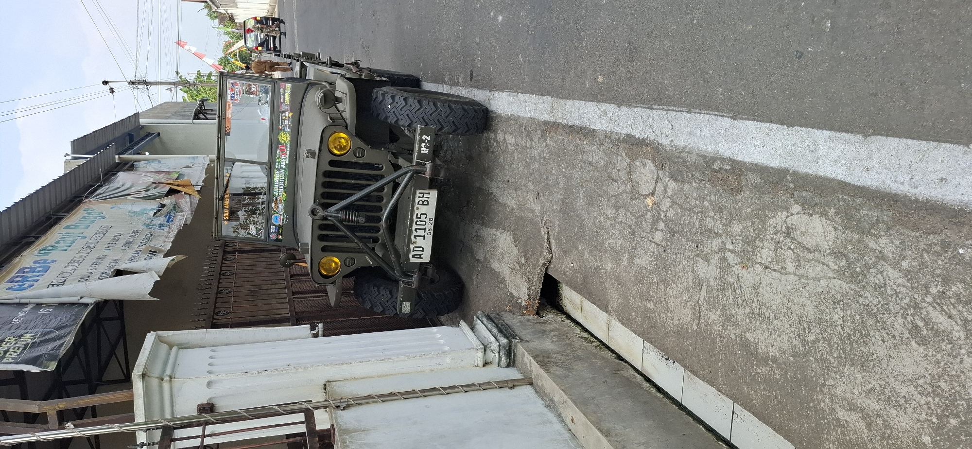 In Gentan, Indonesia, a vintage olive-green Jeep is parked curbside on a paved road, with its front bumper angled towards a concrete drainage ditch. The Jeep features bright yellow headlights, a prominent black metal grille, and a license plate reading "AD 1105 BH" with "05-28" below it. It is adorned with various stickers on its windshield and hood, including one that says "JAMBOREE AMERICAN JEEP". Behind the Jeep, a dark-colored SUV and a person in military-style attire are visible on the road, with a person in a brown uniform walking between them. The backdrop includes a building with signage, a brown metal fence, and utility poles with wires against a cloudy sky. A white pillar and a section of a building with white tiles line the left side of the image, bordering the drainage area.