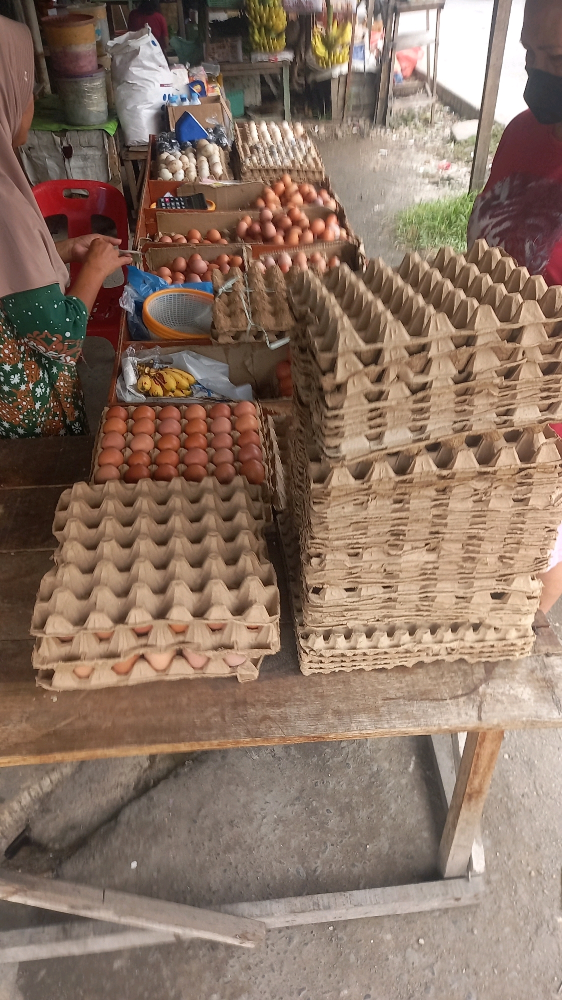 A street market stall in the City of Medan, Indonesia, displays stacks of cardboard egg cartons and trays of eggs. The stall is made of rough wood and sits on a concrete ground. In the foreground, two large stacks of empty egg cartons are visible, one to the left and one to the right, both made of light brown, recycled cardboard. To the left of the empty cartons, a tray filled with light brown eggs is arranged in rows. Behind this, more trays of eggs, some with darker shells, are stacked. In the background, a person wearing a pink headscarf and a green and white patterned top is seen from the side, interacting with the stall. Another person, partially visible on the right, is wearing a red shirt and a black face mask. Various food items, including bananas, are also present at the stall, along with bags of produce and other market goods. The overall scene depicts a busy, outdoor market environment.