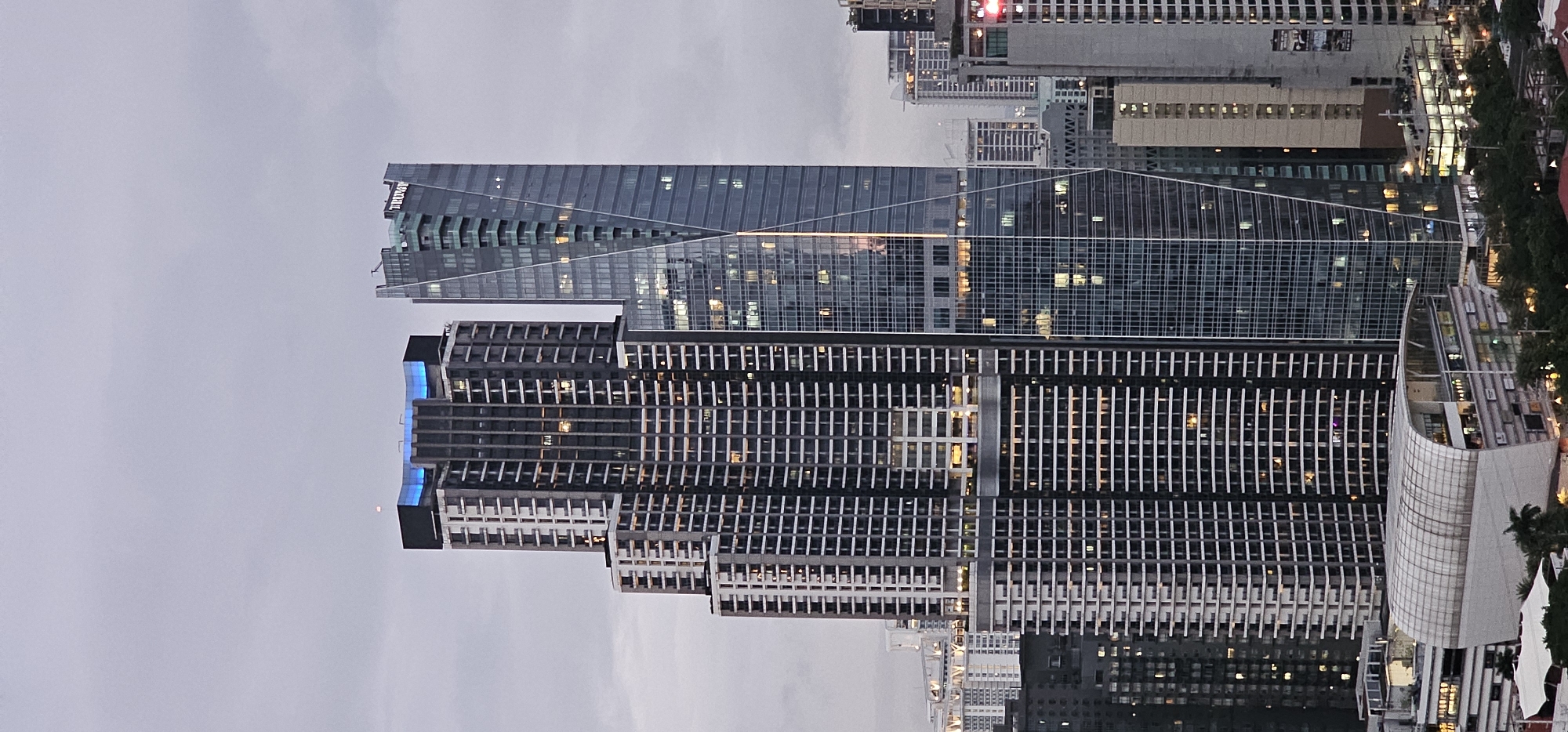 The image shows a cluster of skyscrapers in Makati, Philippines, under an overcast sky. The prominent buildings are a tall, dark, rectangular skyscraper on the left with numerous horizontal white balconies or window frames, and a sleek, glass-clad skyscraper to its right with a unique angled design. The glass skyscraper has the word "TRUMP" illuminated in white letters near its apex. Several windows in both buildings are lit, suggesting occupancy. In the foreground, a modern, curved building with a white, ribbed roof is visible, appearing to be a commercial or retail space. Surrounding these structures are other high-rise buildings of varying heights and architectural styles, some with illuminated windows. The overall impression is a dense urban landscape characterized by modern architecture.