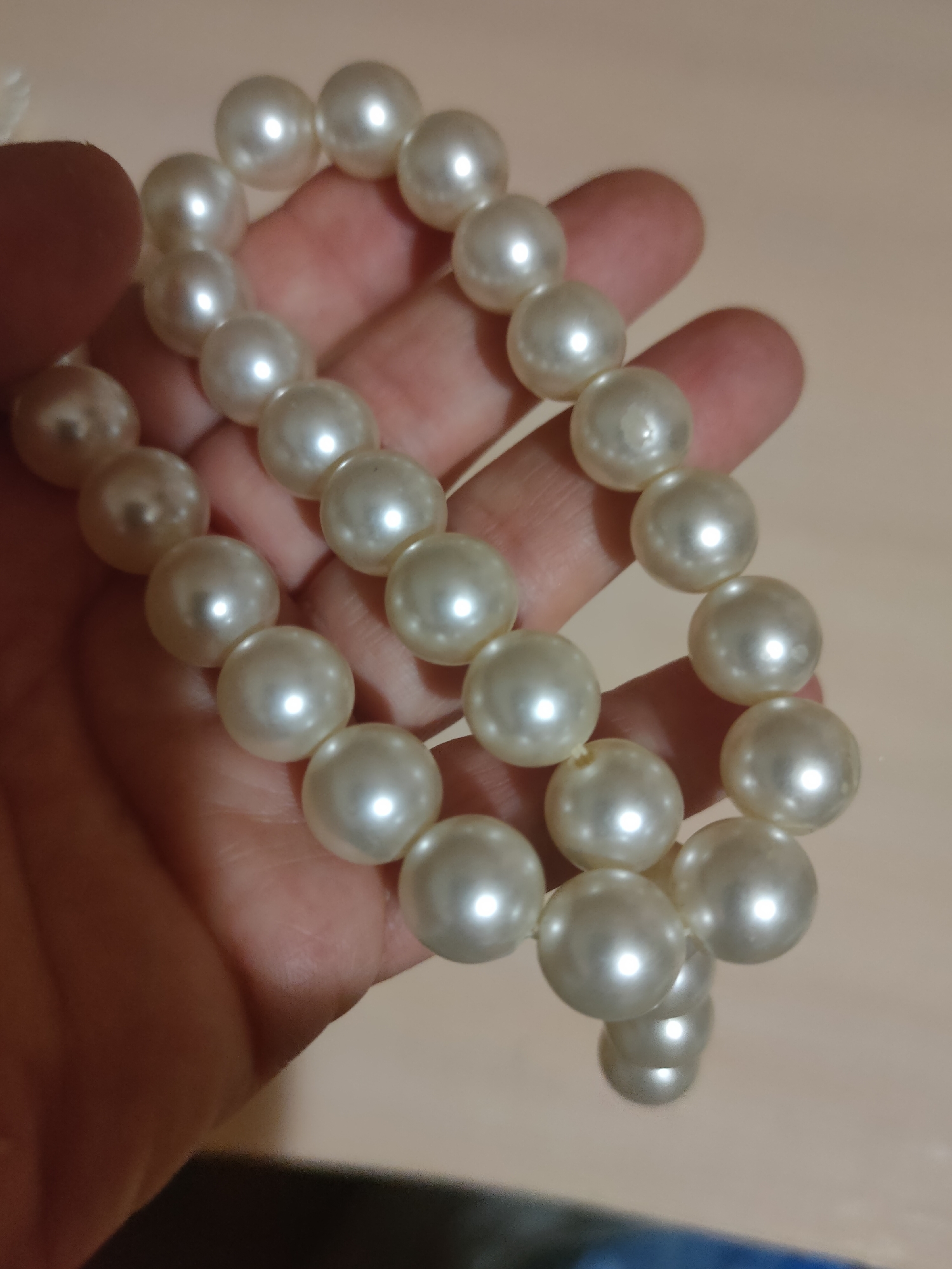 The image displays a close-up of a pearl necklace being held by a person's hand. The pearls are round and white, with a lustrous finish that reflects light. The hand, which appears to be a light-skinned adult, is cupped and gently supporting the necklace, allowing a portion of it to cascade downwards. The background is a soft, neutral tone, suggesting an indoor setting. The lighting is gentle, highlighting the texture and sheen of the pearls. The image does not contain any specific details that would allow for identification of its location within the city of Oktyabrsky, Russia.