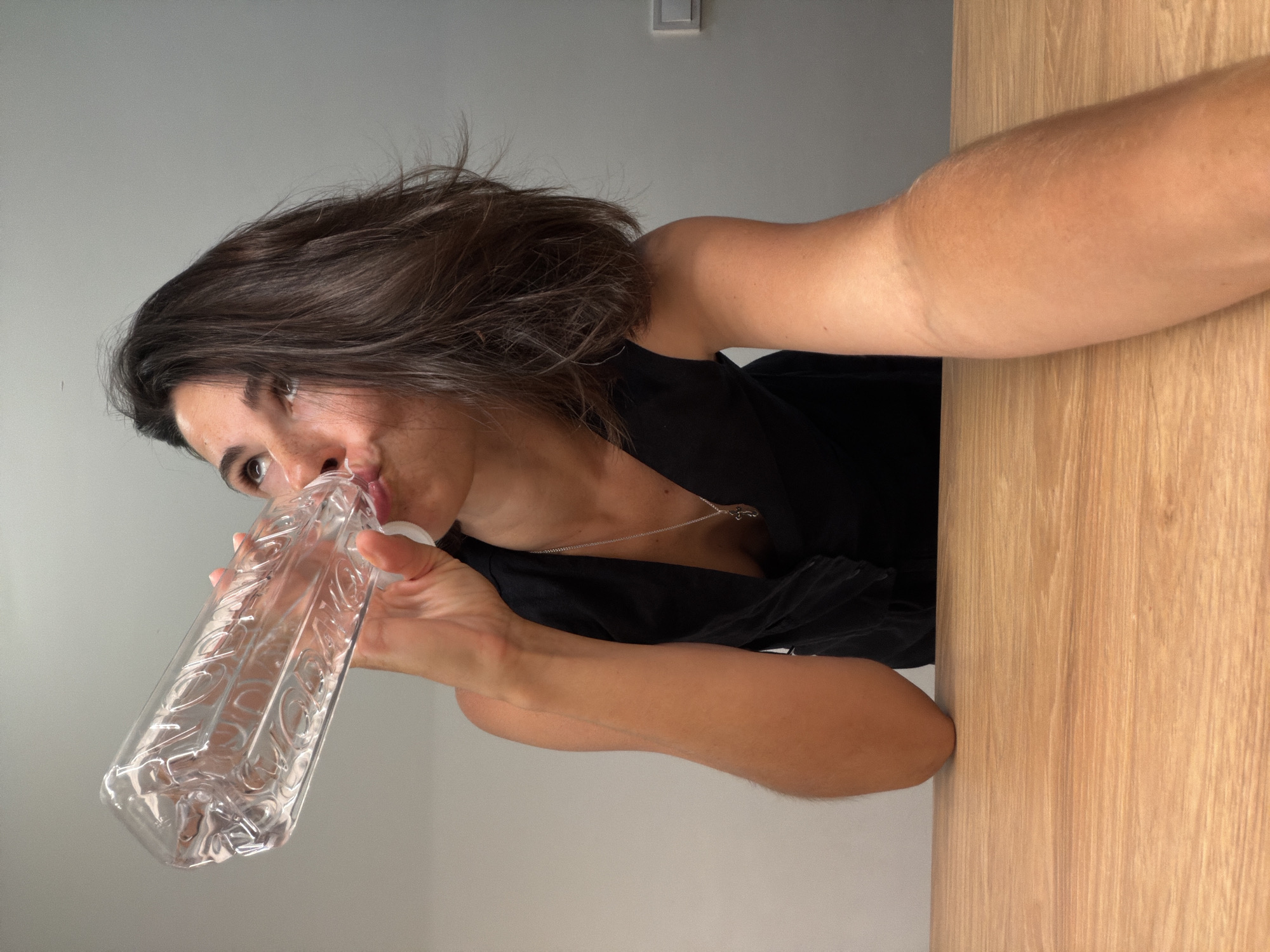 An indoor, portrait-oriented image, rotated 90 degrees clockwise, depicts a woman drinking from a clear plastic bottle. The shot is taken from a low, upward angle, likely a selfie. The woman has medium-length dark brown hair and is wearing a dark, possibly black, top or dress, along with a delicate silver chain necklace. Her mouth is placed on the opening of the rectangular plastic bottle, which features intricate embossed patterns. Her eyes are partially visible, looking slightly upwards.
The background consists of a plain light grey wall. A white light switch is visible on the wall in the upper right section of the frame. To the woman's right, a light-colored wooden surface, possibly a table or a door, occupies the side of the image. Her right arm, extended and partially visible, suggests she is holding the camera. The lighting is bright and even, indicating a daytime setting. The image captures a casual moment of hydration. No readable text is visible on the bottle or in the background.