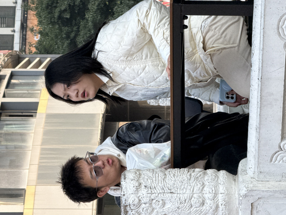 The image depicts two East Asian individuals seated outdoors in Yuzhong District, China. On the left, a male subject with short, dark hair and clear-framed tinted sunglasses wears a white hooded sweatshirt under a black jacket. His mouth is slightly open, and he has a white plastic bag near his chest. To his right, a female subject with long, dark hair wears a white quilted jacket over a light-colored shirt and light beige ribbed pants. She has a wide-eyed, open-mouthed expression suggesting surprise or intense focus. Her left hand rests on a dark-framed rectangular table that separates her from the male subject. Her right hand holds a light blue smartphone horizontally, partially obscured by her thumb. A decorative white stone railing with carved motifs is visible in the immediate foreground on the left. In the background, a multi-story building with a light-colored facade and windows, along with some green foliage, is present.