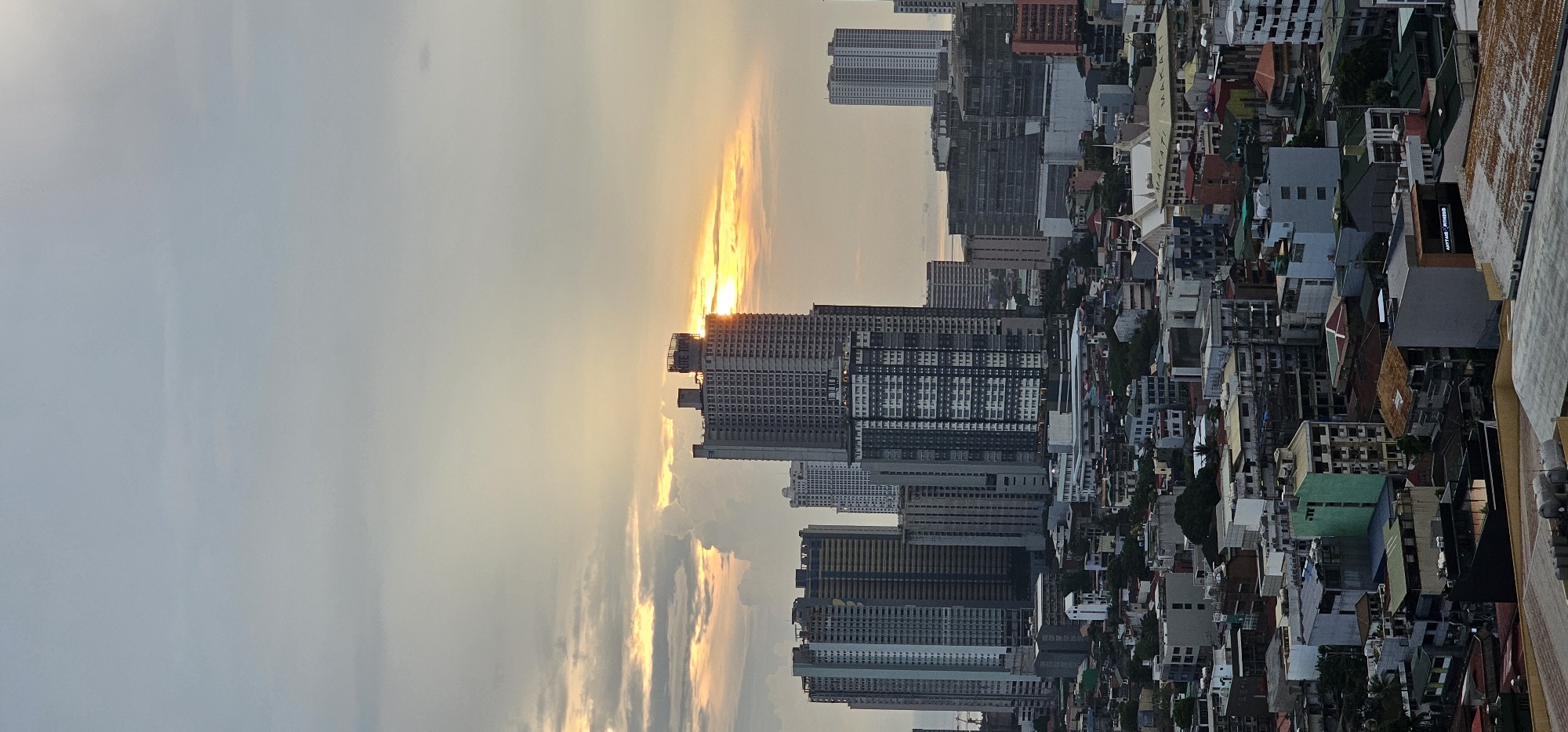 This is a high-angle, vertical shot of a cityscape at sunset. The sun is setting behind a large, modern apartment building, casting a warm orange glow through the clouds. The city is densely populated with a mix of tall skyscrapers and smaller, older buildings. The architecture suggests an urban environment, likely in Asia. The colors are muted by the fading light, with the sky transitioning from a pale blue to a hazy orange and yellow. The buildings are varied in height and design, with many concrete structures, some with visible balconies, and others with flat roofs. The smaller buildings in the foreground appear to be a mix of residential and commercial properties, with some featuring colorful awnings and rooftops. Visible text on one building in the distance reads "MAKATI". The overall mood is peaceful, capturing the transition from day to night in a bustling metropolis. The weather appears to be slightly overcast, with clouds diffusing the sunlight.