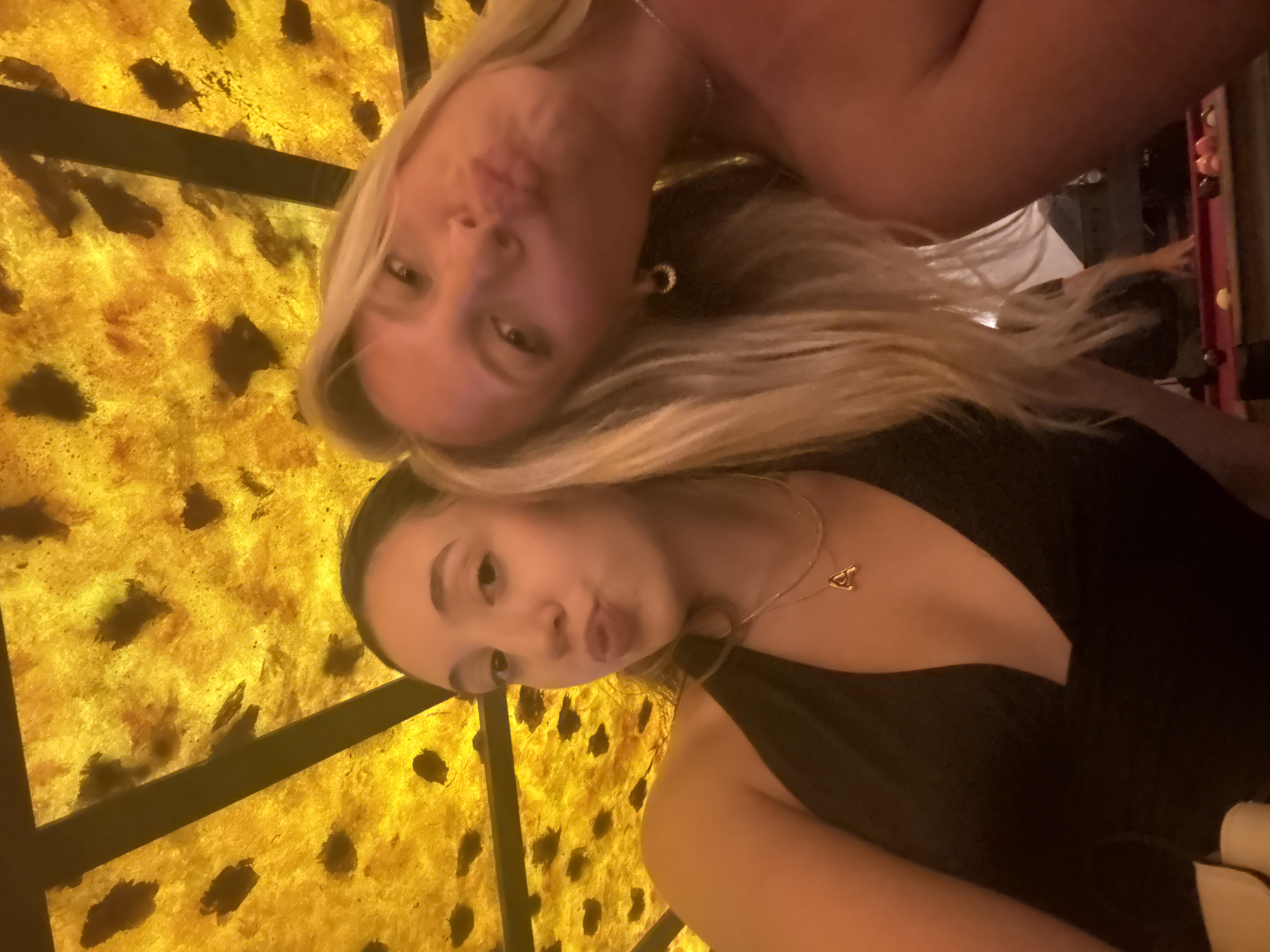 The image captures a close-up, upward-angled selfie of two young women in an indoor establishment in Miami, United States of America. The woman on the left has dark hair and is wearing a black halter-neck top with a layered gold necklace, one strand featuring a small 'A' initial pendant. Her lips are pursed in a pout. The woman on the right has long blonde hair and is partially visible, primarily her face, also displaying a similar pout with pursed lips. She wears small gold hoop earrings and a delicate necklace. Both women are looking directly into the camera. Above them is a distinctive, backlit ceiling composed of a textured, golden-yellow material embedded with numerous irregular dark, organic shapes, resembling dried leaves or abstract patterns. A dark structural beam diagonally crosses the ceiling. In the lower right background, a red-felted billiard table with multiple billiard balls is visible, along with the blurry figure of another person standing nearby.