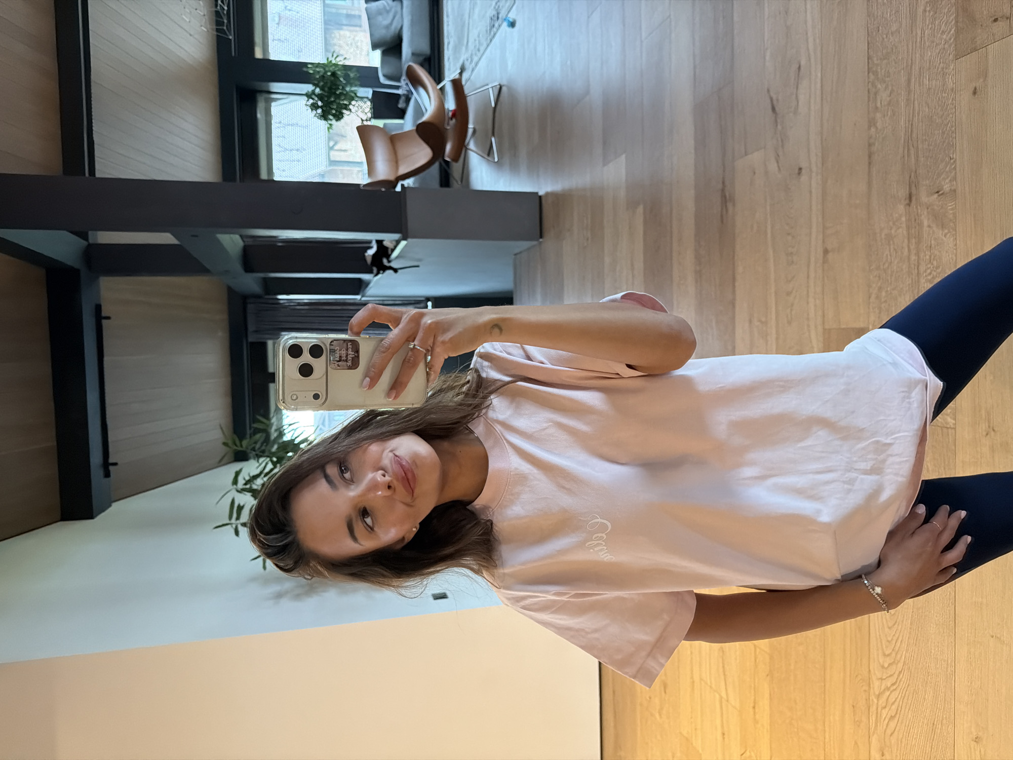 A woman stands in what appears to be a well-lit, modern interior, taking a selfie with her smartphone. The location is not determinable as Shamoni, Russia, based on the visual information provided. She is wearing a light pink t-shirt and dark leggings. Her hair is brown, shoulder-length, and styled with a side part. She is holding her phone in her left hand. Her right hand rests on her hip, and she wears a bracelet on that wrist. The background shows elements of contemporary design, including wood flooring, a black structural beam overhead, and a partial view of furniture. Plants are visible in the background, contributing to the interior décor. A chair with a curved back can be seen in the distance.