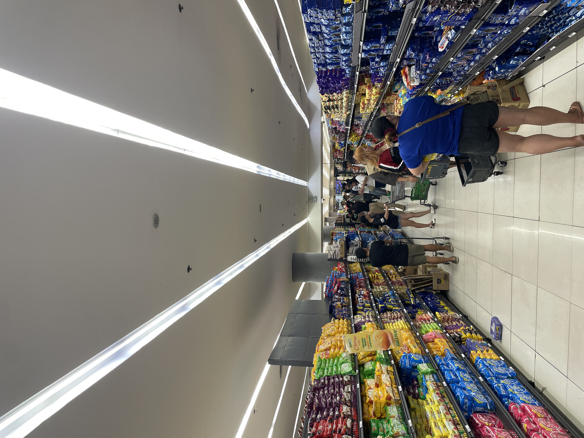 The image depicts the interior of a brightly lit supermarket aisle, likely in Davao City, Philippines, given the product packaging which includes Filipino text. The scene is busy with several shoppers navigating the aisles and interacting with the products on display. The aisle is lined with tall shelves stocked with various packaged goods, predominantly snacks and biscuits. Prominent brands and packaging colors like blue (possibly Oreos) and yellow are visible.
In the foreground, a person wearing a blue t-shirt and black shorts is seen from the back, pushing a shopping cart. To their right, another shopper, a woman in a black shirt and shorts, is also visible. Further down the aisle, more people are browsing and shopping. The floor is tiled, and overhead, long fluorescent light fixtures illuminate the space. The overall impression is that of a typical, active grocery shopping environment.
