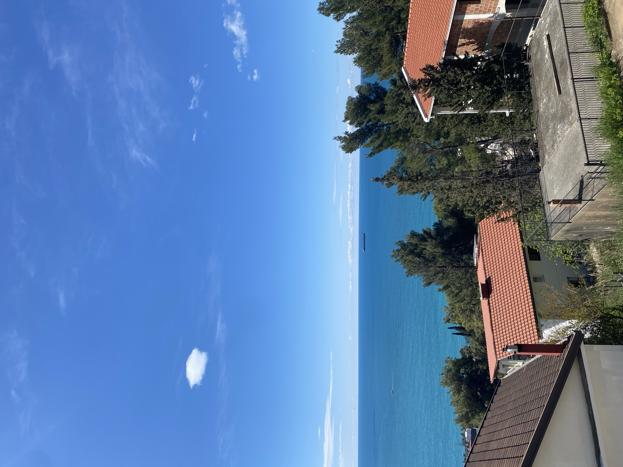 A sunny, daytime view of the Adriatic Sea from Šušanj, Montenegro. The scene includes a clear blue sky with scattered wispy clouds and a single, more substantial cumulus cloud. The sea is a vibrant turquoise, with a distant cargo ship and a small boat visible on the horizon. Foreground details include houses with orange and brown tiled roofs interspersed with dense green trees. A section of a concrete structure with a metal fence runs along the right edge of the frame. The image has been rotated 90 degrees counterclockwise.