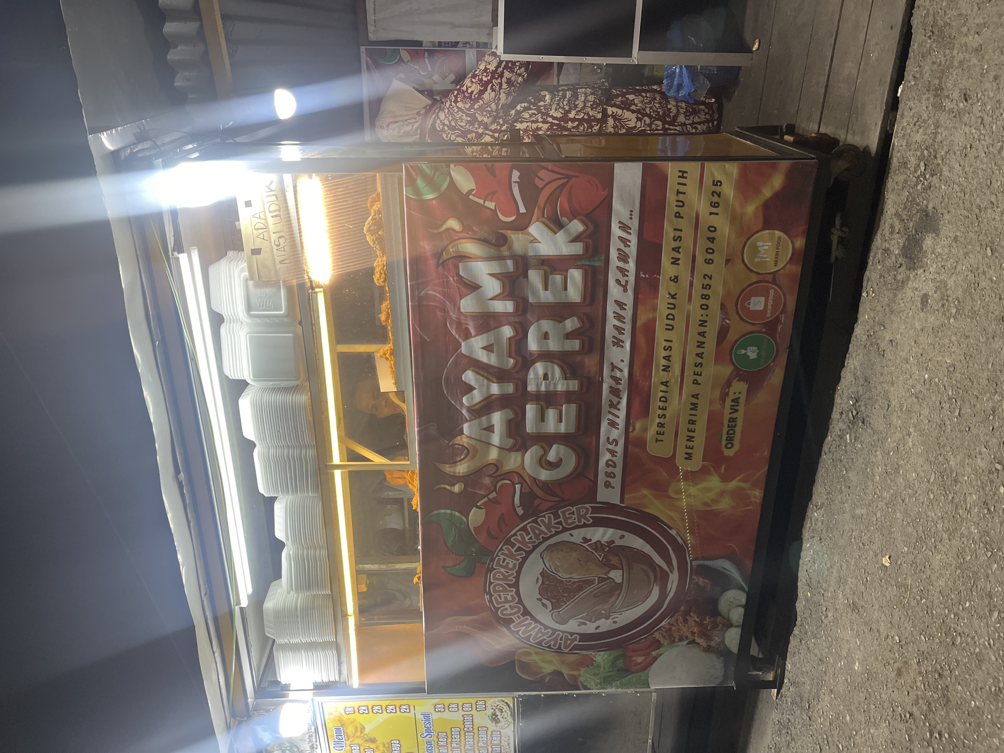 The image shows an outdoor food stall at night, likely in Banda Aceh, Indonesia. The stall is illuminated by bright lights and features a prominent banner advertising "AYAM GEPREK KAKER," which translates to "Smashed Chicken Kaker." The banner also displays the slogan "PEDAS NIKMAT, HANYA LAWAN..." meaning "Deliciously Spicy, Only for the Brave..." and offers "AYAM GEPREK NASI UDUK & NASI PUTIH" (Smashed Chicken with Coconut Rice and White Rice). A phone number, 0852 6040 1625, is provided for orders, along with icons for food delivery services like ShopeeFood and Maxim Food.
Stacked white styrofoam containers are visible on shelves above the counter, ready for food service. A person, partially obscured by the stall and wearing a headscarf and patterned clothing, stands to the right of the stall. The stall itself appears to be constructed from metal and wood. The ground in front of the stall is paved with asphalt, with some debris visible. The overall scene suggests a busy, nocturnal food vending operation.