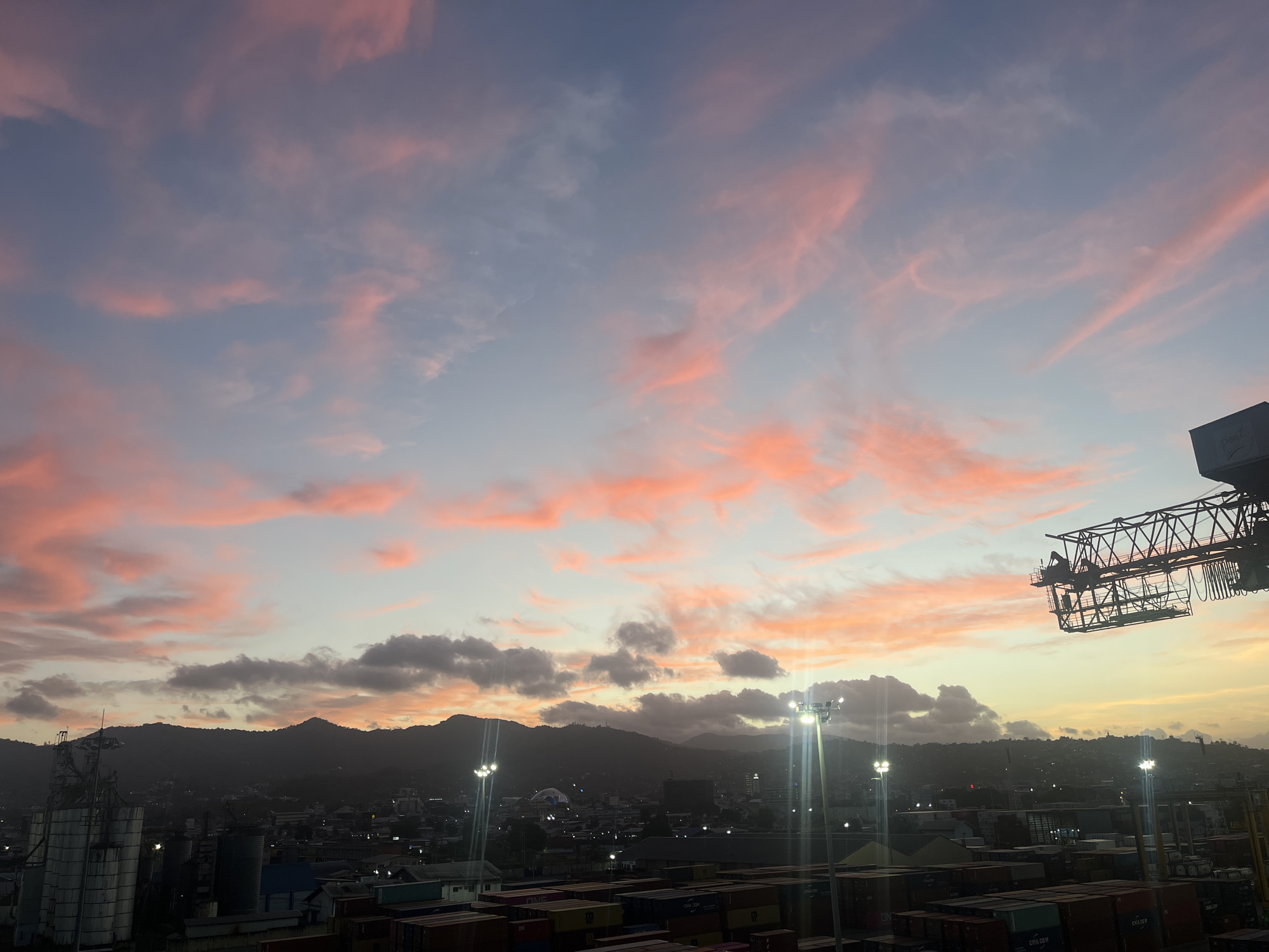 The image presents a panoramic view of an industrial port city at either sunrise or sunset, characterized by a striking sky and a blend of natural and man-made elements. The upper two-thirds of the frame are dominated by a sky featuring wispy, elongated clouds painted in vibrant shades of pink and orange, contrasting with areas of light blue-grey. Closer to the horizon, the clouds appear denser and darker, retaining some of the warm hues.
A range of dark, silhouetted mountains or hills forms a jagged horizon line across the middle of the frame. Below the mountains, the lights of a sprawling city or town are visible, indicating urban development. The foreground is distinctly industrial, featuring a large number of stacked shipping containers of various colors, suggesting a busy cargo port. On the left, tall, cylindrical silos are prominent. Bright, tall floodlights illuminate sections of the industrial area, casting sharp beams into the twilight. A large gantry crane or similar heavy machinery is partially visible on the far right, its intricate metal structure silhouetted against the colorful sky.
The time of day is clearly twilight, marked by the dramatic sky colors and the artificial lights being active. No discernible human activity is taking place within the captured moment. Visible text includes "CMA CGM" on several shipping containers, and "ONE" (Ocean Network Express) on another. A faint logo or text resembling "psl" or "psi" is also visible on a structure in the upper right portion of the image. The scene offers a juxtaposition of the natural beauty of the sky with the industrious nature of a modern port.