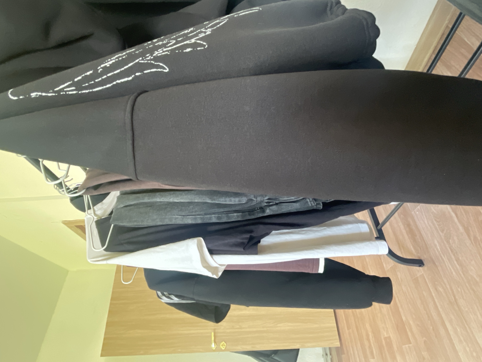 This indoor photograph captures a collection of clothes hanging on a black metal clothes rack in what appears to be a room in Красные Баки, Russia. The room features light green walls and light brown wooden flooring.
The primary subjects are the various garments. A black hoodie with a distinctive white, abstract, or stylized design on its upper part is prominent at the top. Below it, several other pieces of clothing are neatly arranged, including a dark grey item (possibly jeans or a top), a white shirt, a maroon-colored garment, and another black item, partially visible, which appears to have white stripes. Some of these clothes are held by white wire hangers.
In the background, below the clothes rack, a light brown wooden surface with two small golden handles is visible, suggesting a piece of furniture like a chest of drawers or a shelf. The image is taken from a low, tilted perspective, looking up at the clothes rack. There is no specific activity taking place; it's a static scene. The lighting is ambient indoor light. No text is visible on any of the items or in the setting.