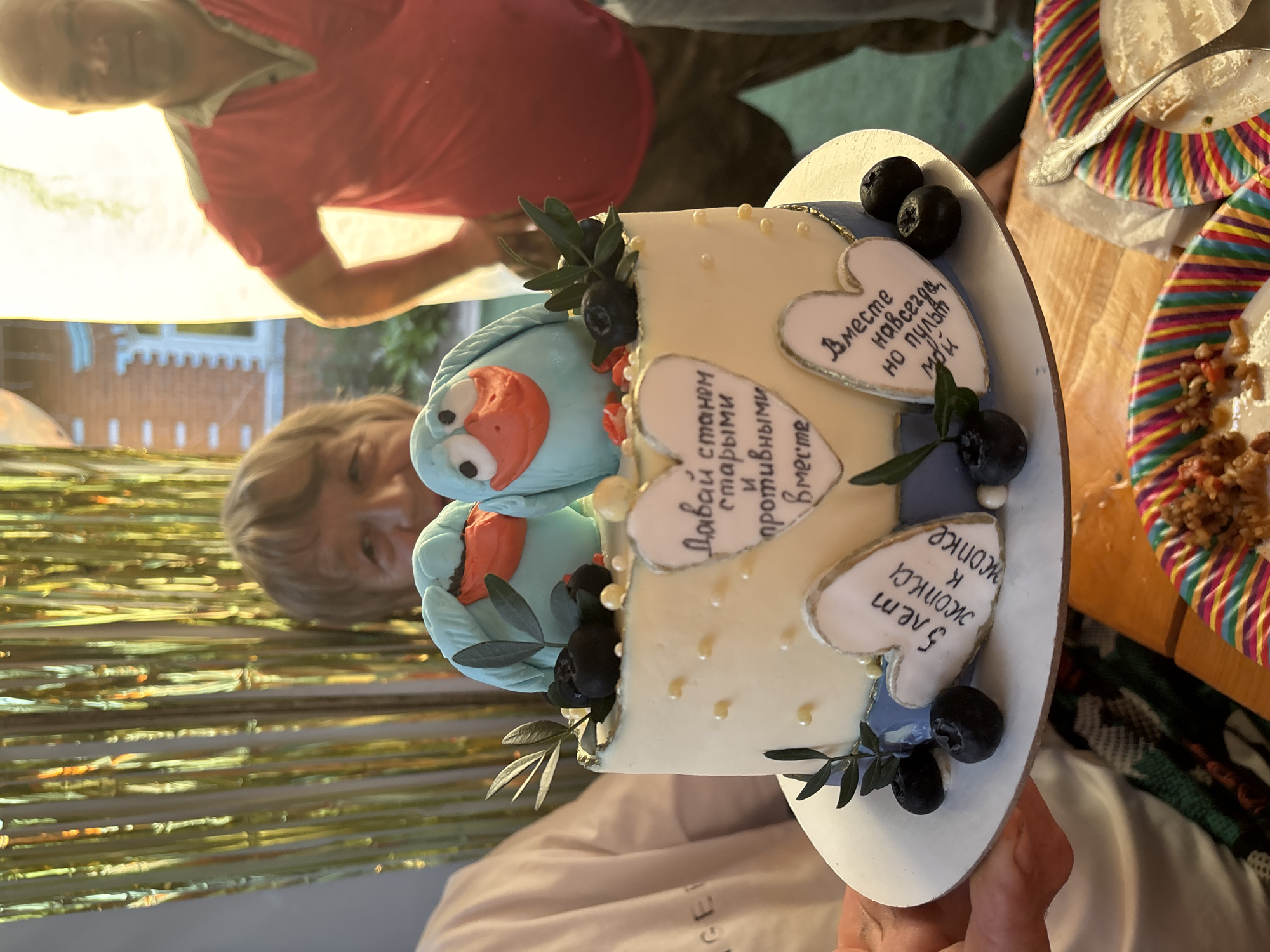 The image depicts a celebratory gathering, potentially an anniversary, in an outdoor or semi-outdoor setting in Rozhdestveno, Russia. A decorated cake is centrally presented, held by two hands with fair skin.
The cake is round, approximately two tiers, with white or light cream frosting. The top of the cake features two light-blue bird figurines with orange beaks and white eyes, facing each other. Edible decorations include dark blueberries and green leaf sprigs. A blue ribbon or band encircles the base of the top tier. Three heart-shaped plaques with gold trim are attached to the cake's side. The central top heart displays Russian text translating to "Let's grow old and nasty together." The bottom left heart reads, "5 years butt to butt." The bottom right heart states, "Together forever, but the remotes are mine." Small white and yellow pearl-like edible spheres are scattered on the frosting.
Behind the cake, an adult female with fair skin, short light-brown hair, and a visible smile is partially obscured, looking directly at the camera. To the right, an adult male with fair skin and short or bald hair is standing, partially silhouetted by bright light. He wears a red polo shirt with a light-colored collar and has his hands on his hips. The background on the left features vertical gold metallic streamers. On the right, a brick building with white window trim is visible, suggesting an exterior or patio location.
In the foreground, a wooden table is visible. Two colorful paper plates with rainbow stripes are present; one contains remnants of a granular, sauced food, while the other is mostly empty with a silver spoon resting on it.