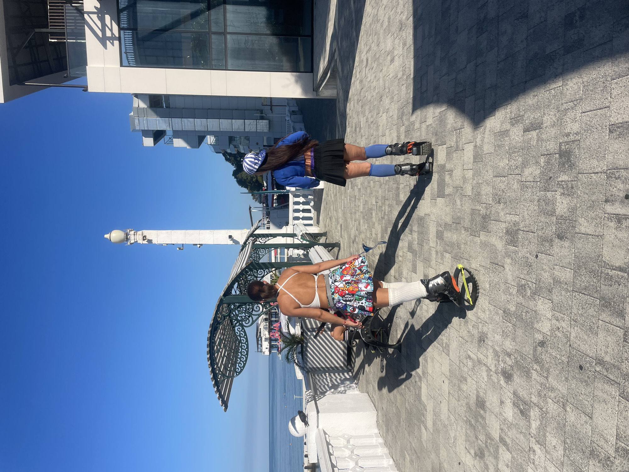 Two women are seen from behind, walking on a paved promenade near the Black Sea in Sochi, Russia. The woman in the foreground wears a white sports bra, a colorful skirt adorned with cartoon characters, and white knee-high socks with jumping shoes. The woman behind her is dressed in a blue long-sleeved shirt, a black mini-skirt, blue knee-high socks, and similar jumping shoes, and is wearing a blue and white striped cap. The promenade features a white railing and benches. In the background, there is a white structure with arched eaves, a tall white tower, a modern white building with glass windows, and the blue sea under a clear sky.