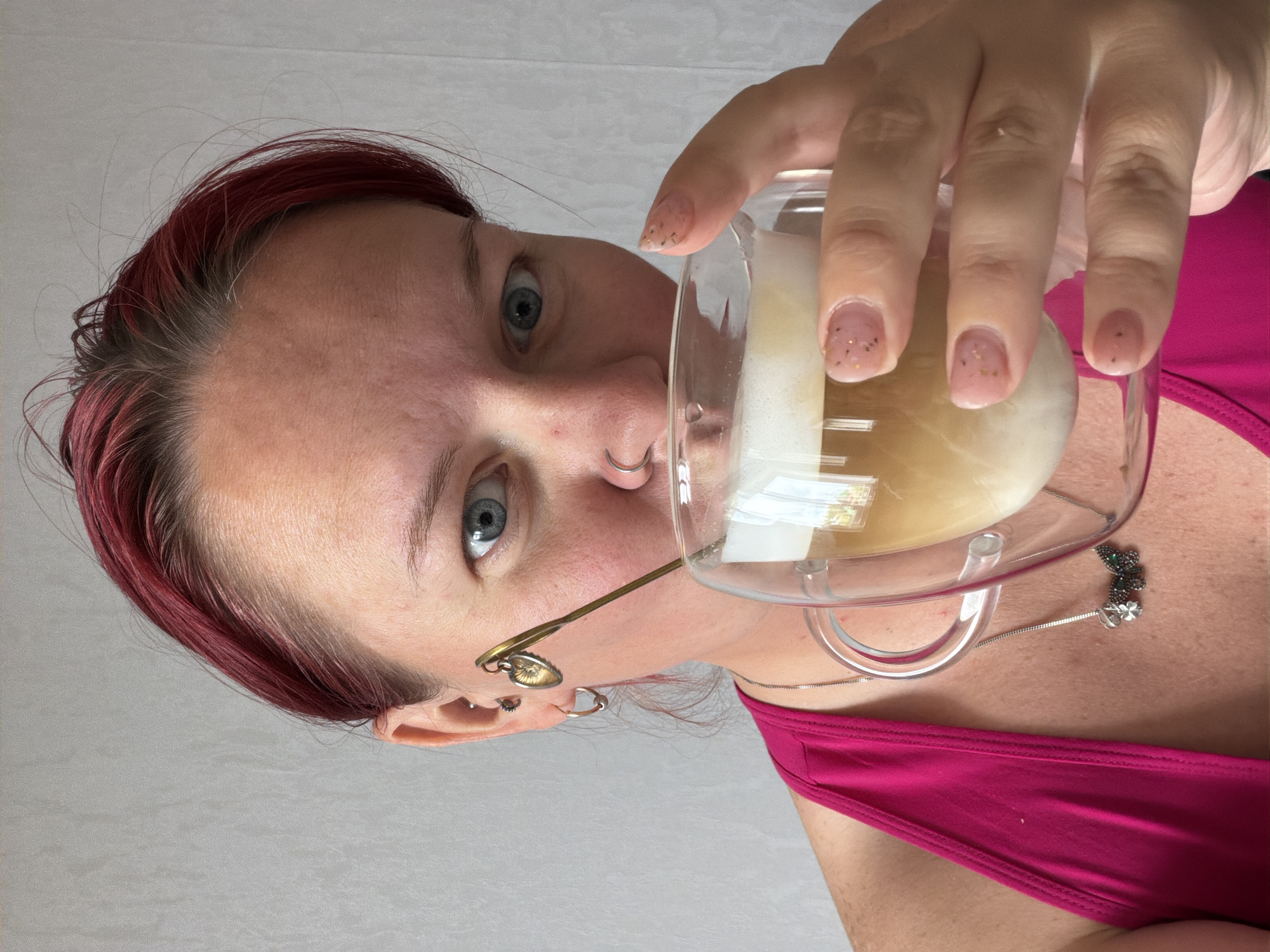 A close-up, high-angle shot captures a person looking directly at the camera while holding a glass with a drink. The image is rotated 90 degrees clockwise, showing the subject's face from a worm's-eye view.
The main subject is a woman with striking blue eyes and auburn or dark red hair, some strands showing natural brown roots and lighter, wispy hairs around the hairline. Her skin appears fair with some freckles or sun spots visible on her forehead. She has a nose ring on her left nostril. Her left ear is visible, adorned with multiple piercings including a small stud and a hoop, and a unique pair of glasses with a brass-colored frame that features a small heart-shaped charm dangling near her ear.
She holds a clear glass mug containing a creamy, light brown beverage, possibly a coffee or latte, with a layer of foam on top. Her nails are manicured with a light pink or nude polish and tiny golden glitter or confetti embedded in them. She is wearing a bright fuchsia or hot pink tank top or camisole. A silver chain necklace with a small, detailed pendant resembling a clover or floral design is visible around her neck, resting on her chest.
The background is a plain, light-colored surface, possibly a wall or ceiling, contributing to the intimate and focused nature of the shot. No text is visible in the image. The overall impression is casual and personal, with the subject engaged in a simple act of drinking.