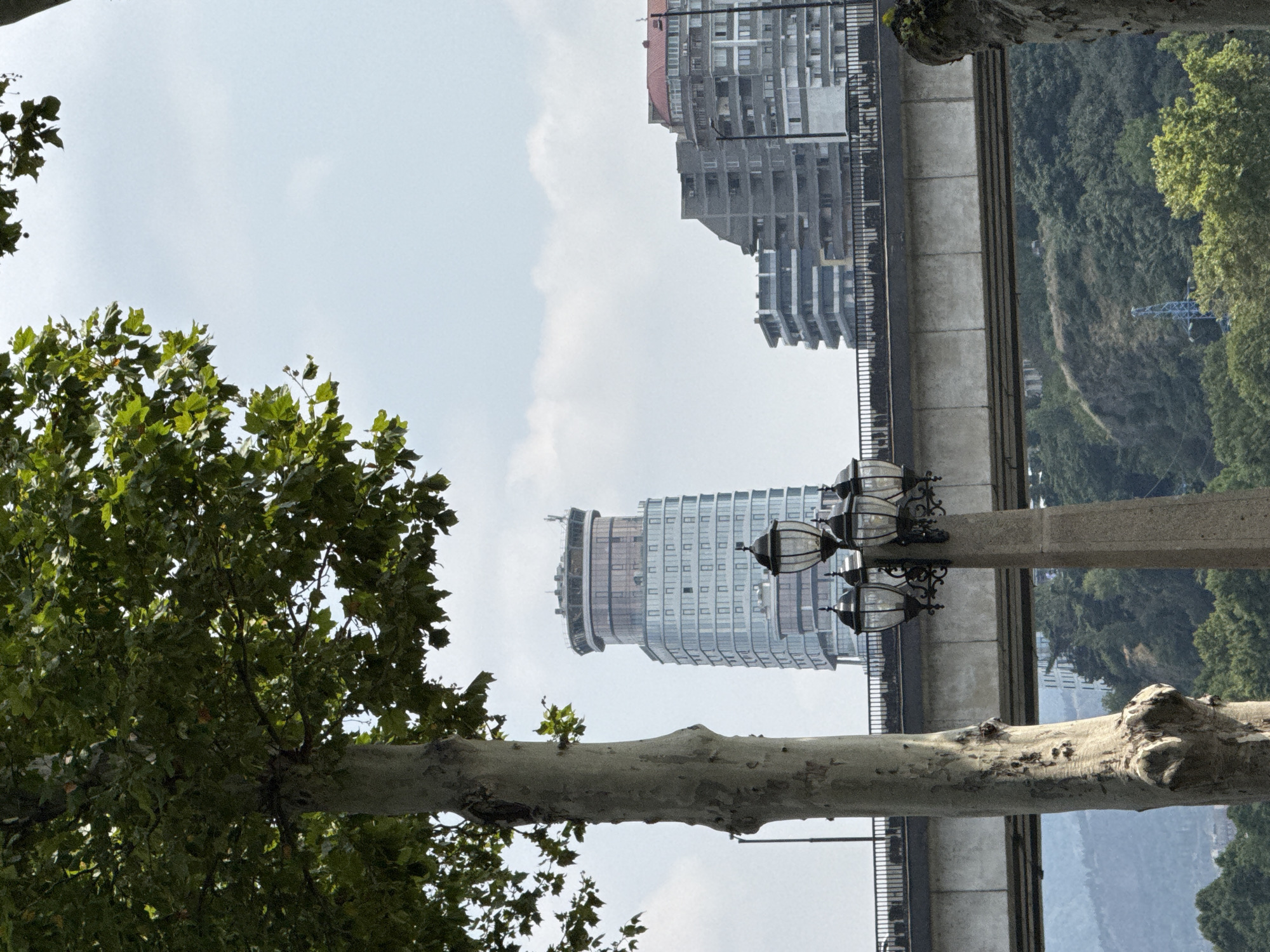 The image shows a street lamp in the foreground, with a tree to its left and a bridge in the background. Behind the bridge, there are several modern buildings, including a tall, cylindrical skyscraper with a glass facade. The scene appears to be in an urban environment, likely in Tbilisi, Georgia, given the architectural style of the buildings. The sky is overcast with some clouds. There are no people visible in the image.
