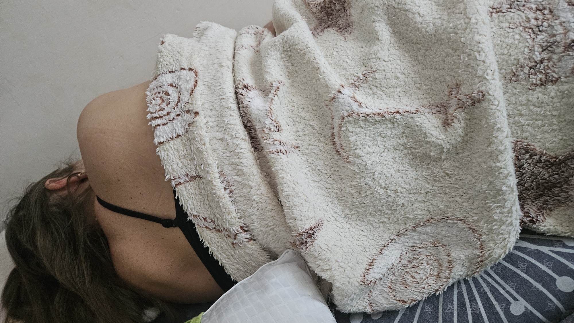A person is lying on their side, facing away from the camera, covered by a fluffy blanket with a rose pattern. The person is wearing a black bra. The blanket is draped over their shoulder and chest, obscuring most of their body. A white pillow is visible to the left of the person. The background consists of a plain, light-colored wall and a patterned duvet. The scene appears to be indoors, likely in a bedroom. The image is cropped tightly on the person and blanket, providing a close-up view. There is no visible indication of the person's location in Bratislava, Slovakia.