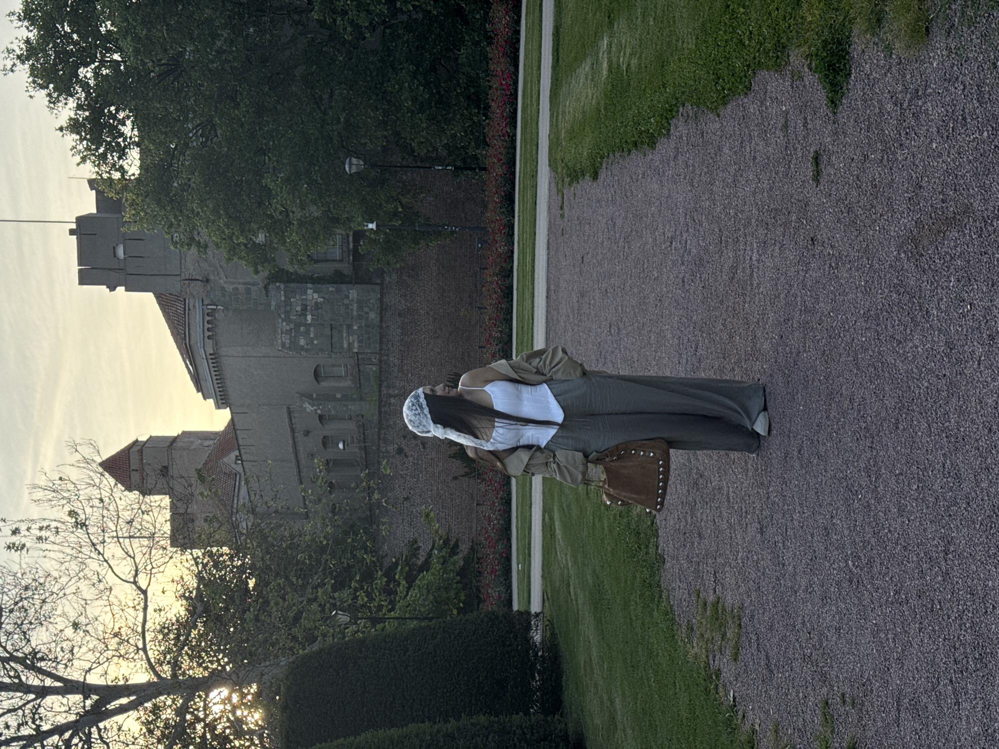 A full-length shot captures a person standing on a gravel path with a castle-like structure in the background. The person, possibly a woman, wears a white top, green pants, a jacket, and a white bonnet. She carries a brown bag on her shoulder. Behind her is a stone building with multiple tiers, resembling a fortress or castle. Lush greenery, including trees and hedges, surrounds the path and the building. The sun is setting, casting a warm glow on the sky and illuminating the trees. The location may be Belgrade, Serbia, but this cannot be verified.