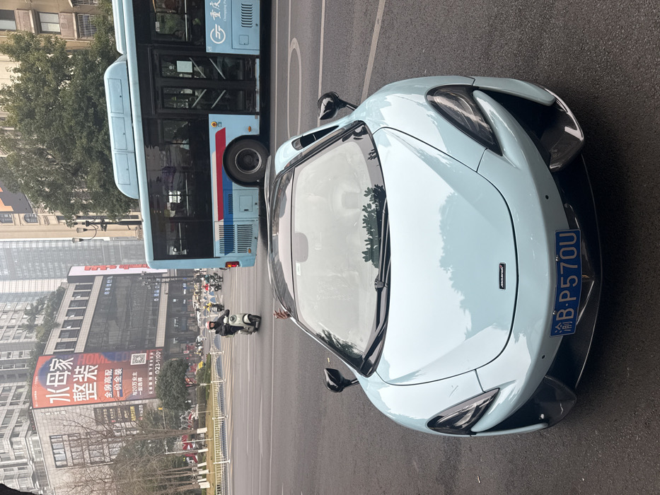 A light blue McLaren supercar is parked next to a blue and white city bus. The car’s license plate reads "渝B-P570U." Buildings and trees line the background. A large advertisement for "HOMESGO" or "水母家" is visible on a building, promoting "全房高配, 一价全装" (high-end full house, all-inclusive price). Scooters and motorcycles are seen on the street further down. The scene appears to be in an urban setting.