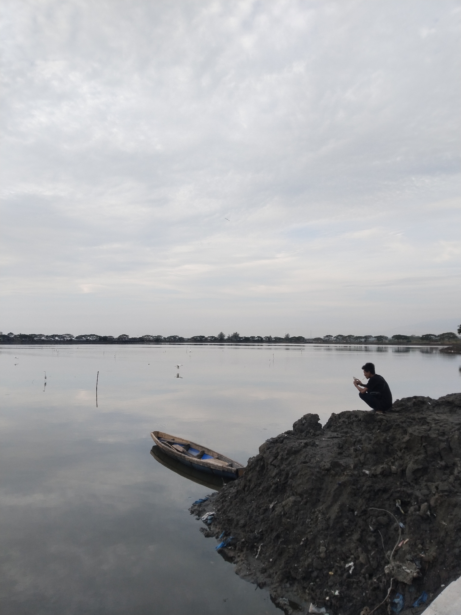 The image depicts a serene, overcast day at a body of water, likely a lake or a shallow sea, with a distant treeline visible on the horizon. A lone person, dressed in a black outfit, is crouched on a dark, muddy embankment, engrossed in their mobile phone. The individual's posture suggests a moment of quiet contemplation or engagement with their device.
In the foreground, a small, weathered wooden boat rests near the muddy shore, its reflection shimmering on the still water. The water's surface is calm, mirroring the cloudy sky and the surrounding environment. A few thin poles or markers emerge from the water, possibly indicating fishing spots or navigational aids. Birds, appearing as small white specks against the vastness of the water and sky, can be seen in the distance, adding a sense of natural life to the scene.
The overall atmosphere is tranquil and somewhat melancholic, amplified by the muted colors and the overcast sky, suggesting it is either early morning or late afternoon. The location, identified as Lhokseumawe, Indonesia, provides a geographical context for the landscape, which appears to be a coastal or lakeside area. The embankment itself is rough and appears to have some debris scattered on it, hinting at a less manicured, perhaps more natural or working-class area. There is no visible text in the image.