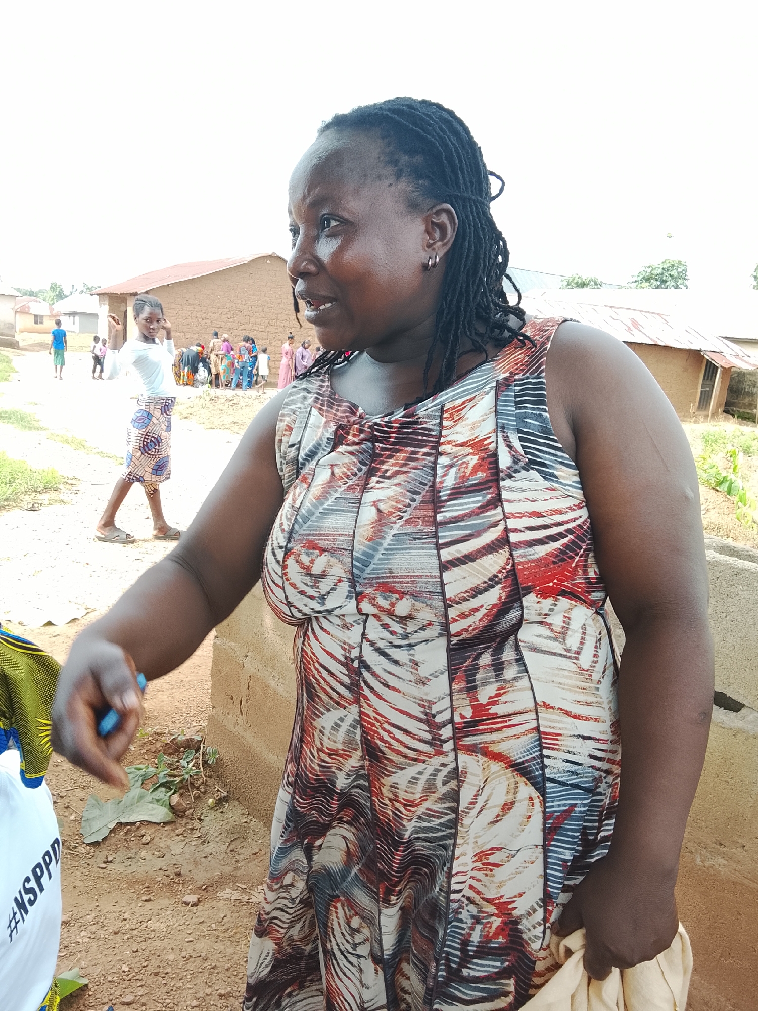 In Akwanga, Nigeria, a woman with dreadlocks and hoop earrings stands outdoors, facing right and looking off-camera. She wears a sleeveless dress with a multi-colored abstract pattern featuring red, white, and black. Her left arm is bent, and her hand is extended towards a white t-shirt with the hashtag "#NSPPD" visible. Behind her, several other individuals, including children, are gathered in front of buildings constructed with mud bricks and corrugated metal roofs. The ground is dirt, and vegetation is sparse. The overall scene appears to be a residential area in a rural or semi-rural setting.