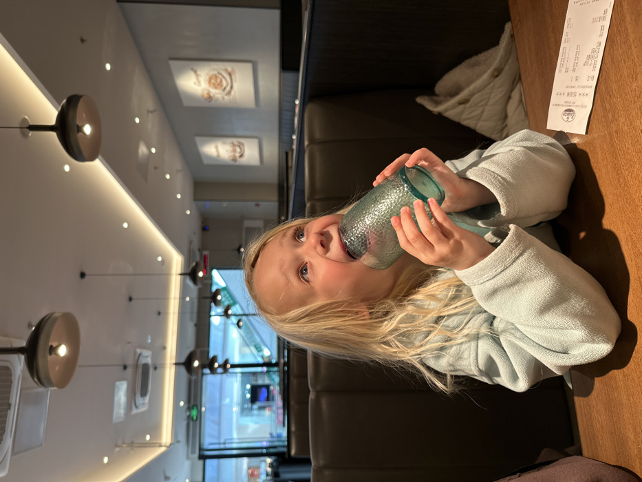 A young girl with blonde hair drinks from a blue textured glass at a restaurant. She wears a light blue sweater. The restaurant has modern pendant lights and artwork on the walls. Outside the window, a commercial area is visible. A dark booth seat and wooden table are in the background. A receipt sits on the table near the girl. The photo is rotated 90 degrees counterclockwise.