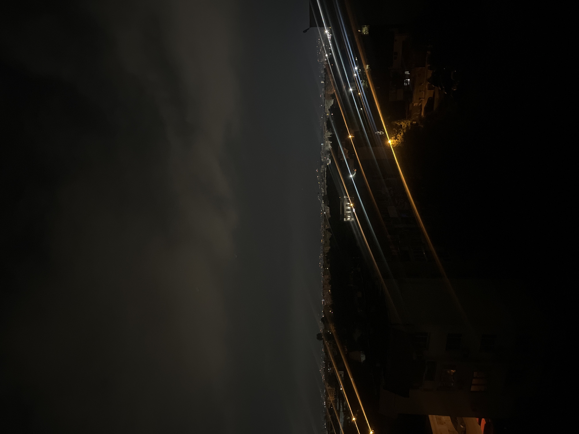 The image depicts a nighttime cityscape in Sofia, Bulgaria. The view appears to be from an elevated position, possibly a high-rise building. The right side of the image is dominated by dark buildings and shadows, with occasional glimpses of illuminated windows. Streaks of light suggest streetlights and moving traffic. The left side features a cloudy or hazy sky with a faint crescent moon visible. The scene is generally dark and illuminated by artificial light sources.