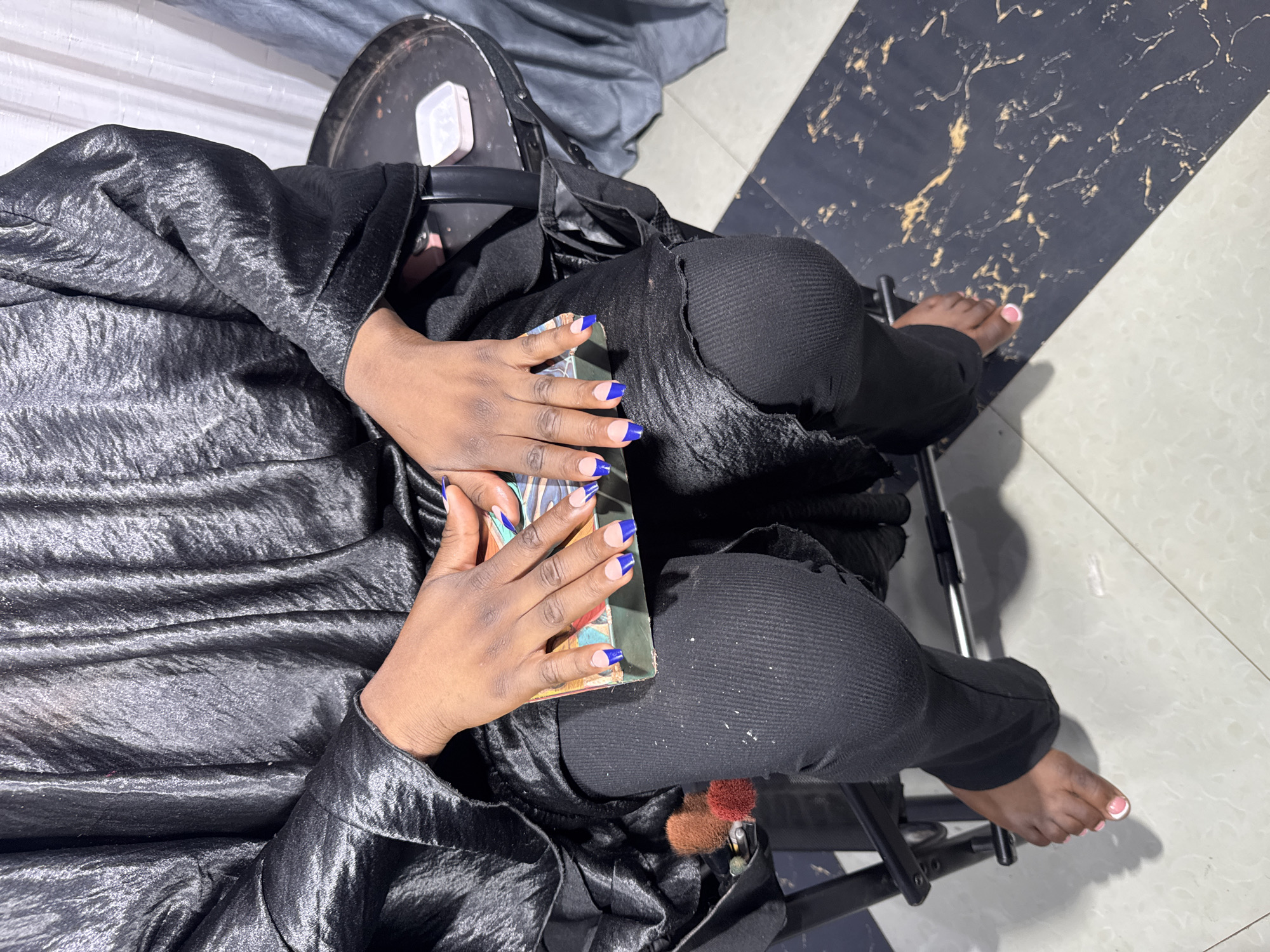 The image shows a person from a top-down perspective, sitting with their legs crossed and wearing a shiny black outfit. The person has dark skin and their nails are painted with a French manicure style, featuring blue tips on their fingernails and pink on their toenails. They are holding a small, rectangular object with colorful patterns, possibly a book or a compact case.
The setting appears to be indoors, with a floor composed of black and white tiles, some with a marbled pattern and gold veining. A dark-colored chair is visible beneath the person, with metal legs and a black seat. In the background, a greyish fabric is draped, and a dark, rounded object, possibly part of a chair or equipment, is also present. The lighting suggests it is daytime. The person's posture and the way they are holding the object imply a moment of repose or engagement with the item. The overall mood is calm and focused on the individual. There is no visible text in the image. The location context provided is Gura Top, Nigeria.