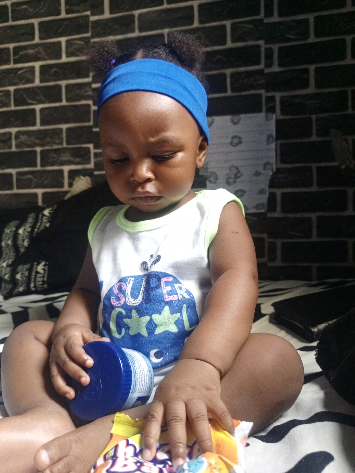A baby is pictured sitting upright, cross-legged, on a patterned surface indoors. The baby has a dark complexion and dark hair styled in two small puffs on the top of the head, secured with a bright blue headband. The baby is wearing a white, sleeveless top with light green trim around the neck and armholes. The top features a graphic that reads "SUPER" in pink and blue letters, followed by a large blue circular design with green stars and a partial 'C' visible, likely part of a word like "CUTE" or "COOL".
The baby's head is tilted down, and the eyes are focused on objects in their hands. The left hand holds a blue cylindrical container with a matching blue lid. The right hand rests on a colorful, partially opened snack bag, which is primarily orange and yellow with blue and white accents and some white text, where "B_B" is legible.
The background consists of a dark wall with a brick-like pattern. The baby is seated on a surface covered by a white and black striped or patterned sheet or blanket. To the baby's left, a dark pillow or cushion with a white pattern is visible. To the right, a dark, flat object, possibly a wallet or phone, rests on the patterned surface. The baby appears focused on the items in their hands.
