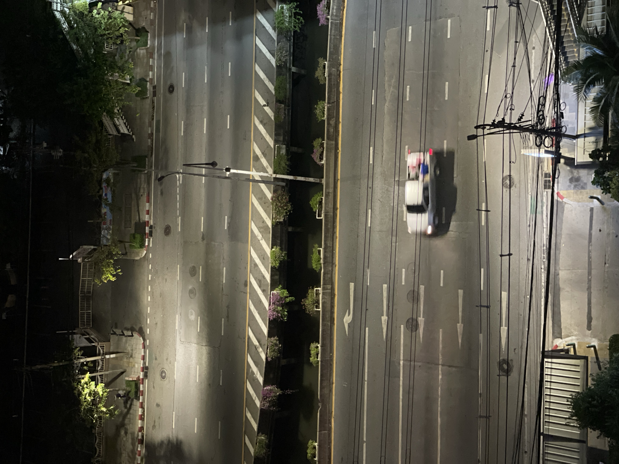 Aerial view of a city street in Bangkok, Thailand at night. The street has multiple lanes of traffic separated by a median with planters and a water feature. A white vehicle is traveling on the right side of the road. Utility poles and wires are visible on the right side of the image. Trees and buildings line the sides of the street. The street is illuminated by streetlights.
