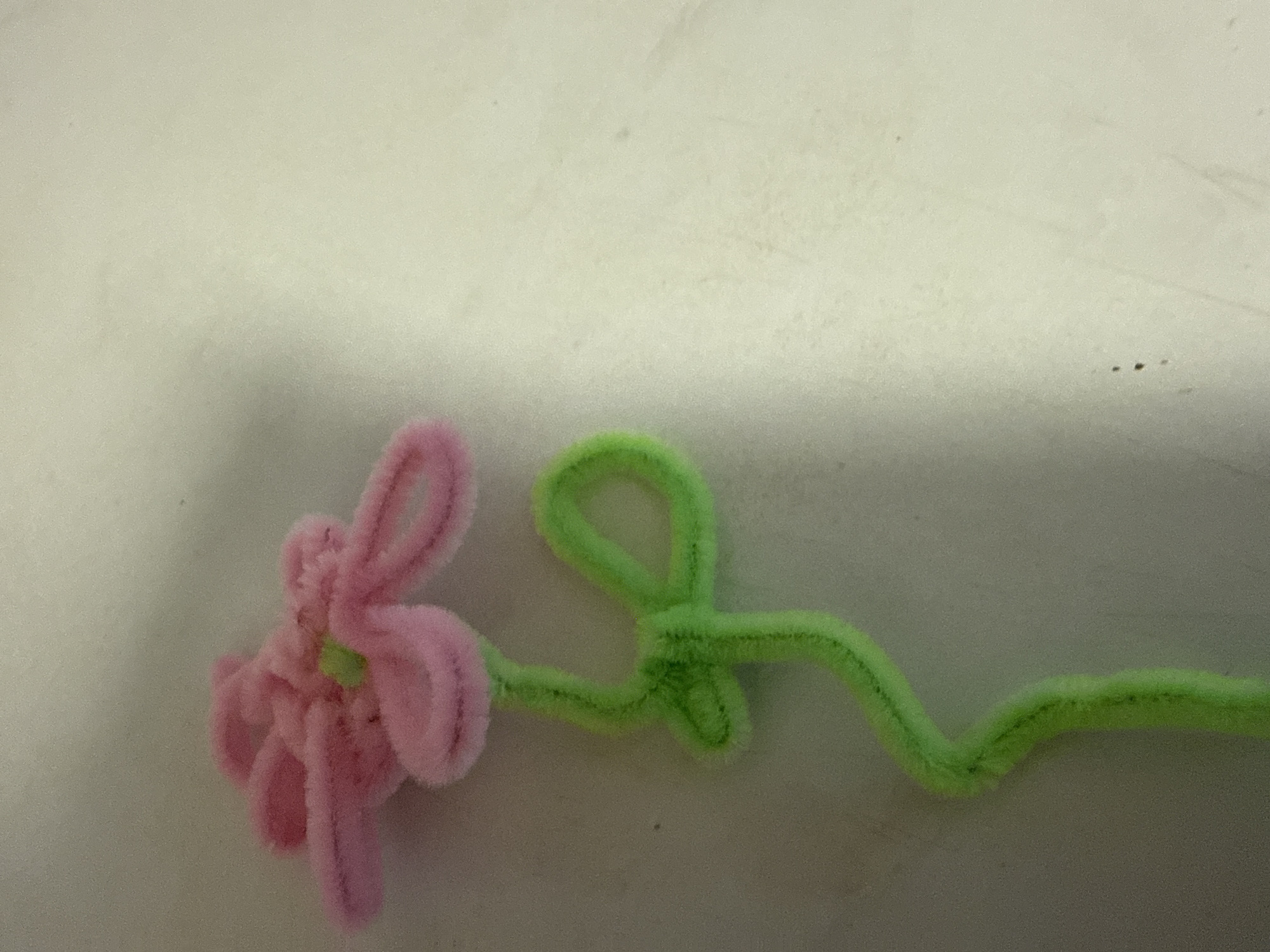 A pink and green pipe cleaner flower is positioned diagonally on a textured, light-colored surface, with the flower's bloom at the top left and its stem extending towards the bottom right. The flower is constructed from fuzzy pipe cleaners, with pink forming the petals and a small green piece at the center. The stem is green and has a curved loop. The background is a plain, slightly mottled surface, possibly a table or wall. The image has a grainy quality and is lit from above. The location is specified as Ridabu, Norway.