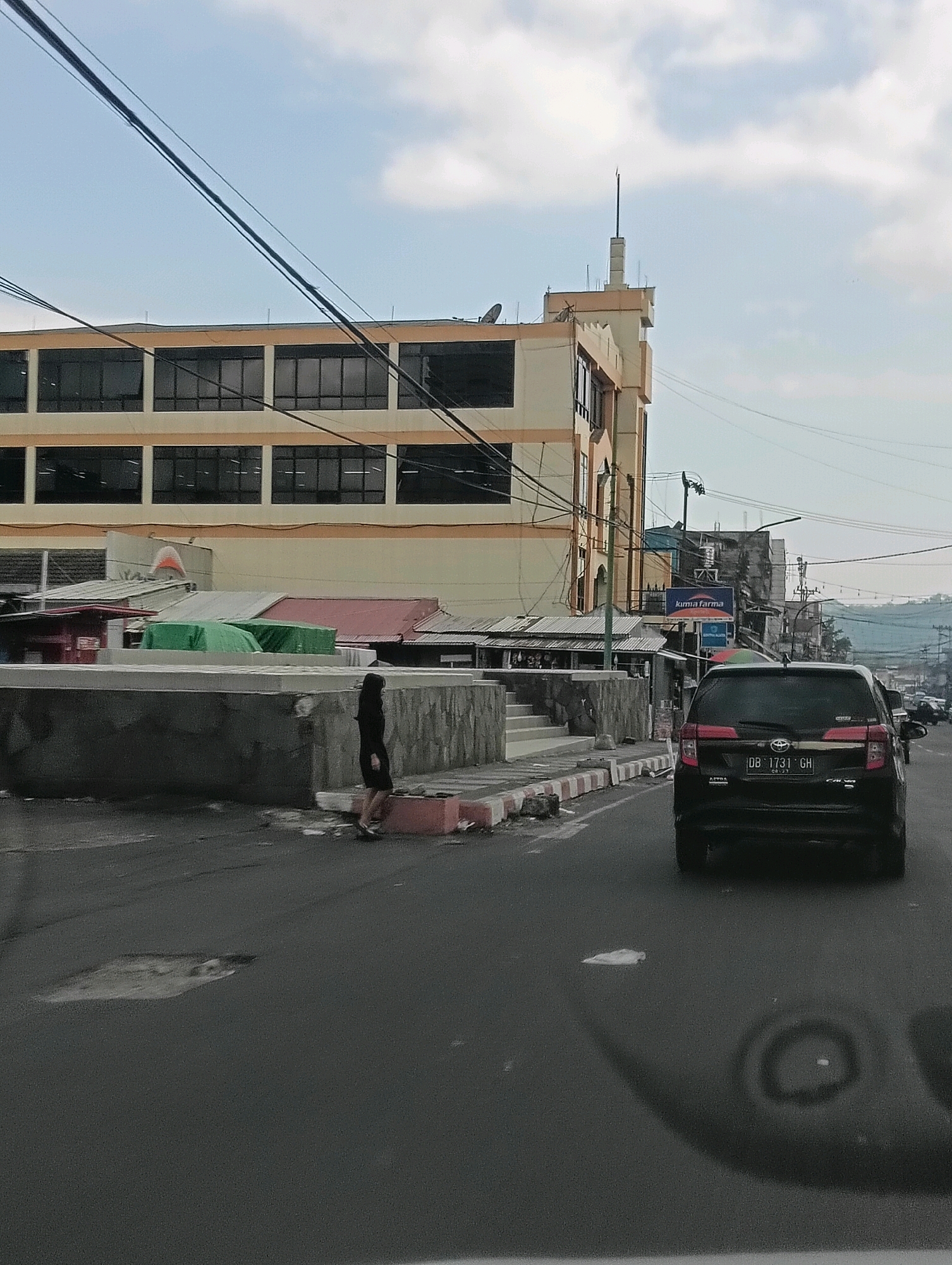 The image shows a street scene in Tomohon, Indonesia. In the foreground, a black SUV is driving down the road. The SUV has a license plate that reads "DB 1731 GH". Further down the road, there is a woman in a black dress walking on the sidewalk. Behind the woman, there is a multi-story building with large windows. The building appears to be a commercial establishment, possibly a department store or office building. There are also several smaller shops and stalls along the side of the road. The sky is overcast with some clouds. The overall impression is of a busy urban environment with a mix of commercial and residential buildings.