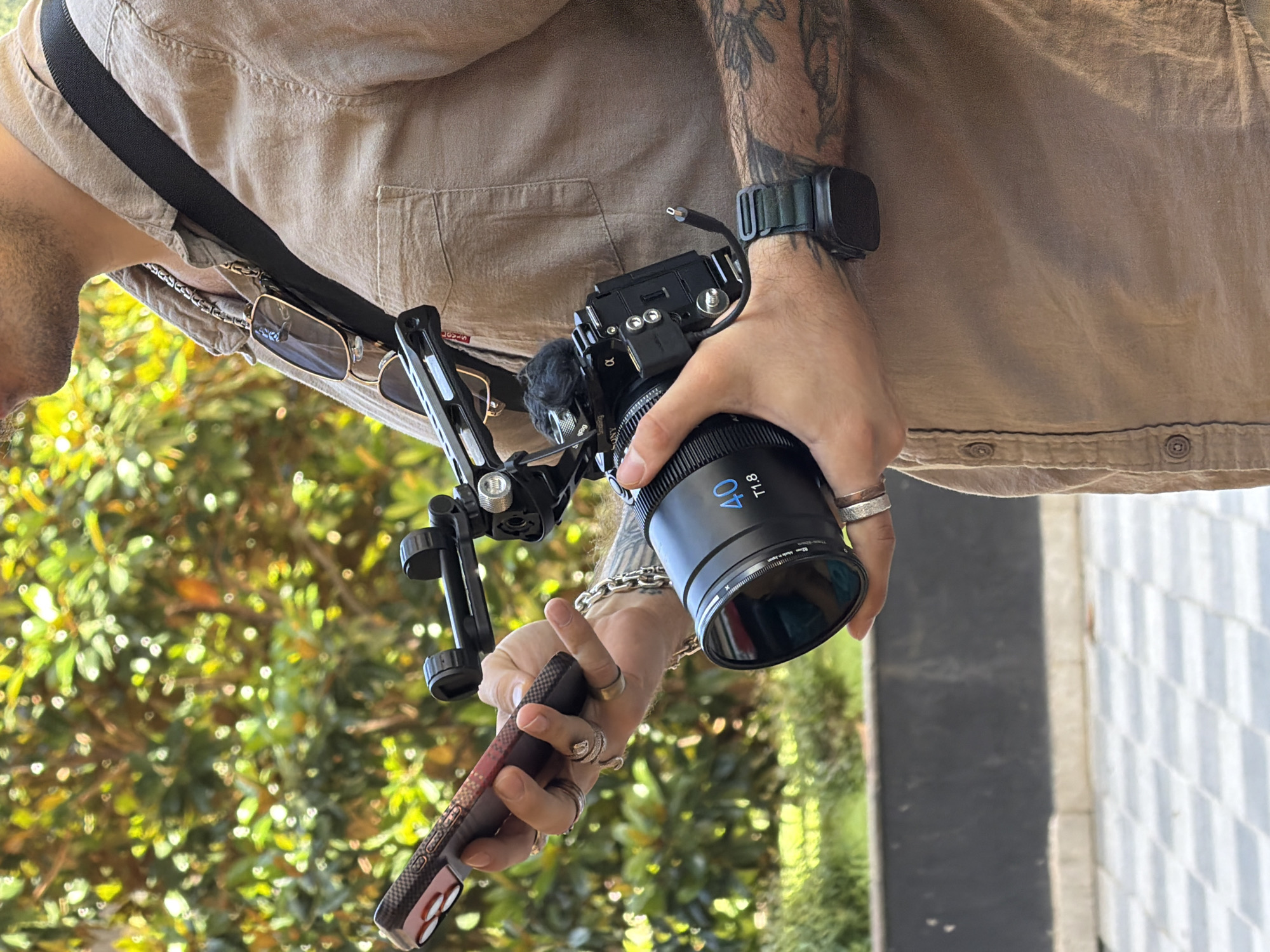 A male subject is depicted outdoors in Budva, Montenegro, shown from the mid-chest upwards, holding a smartphone in his left hand and a camera rig in his right. He wears a light brown button-up shirt with a dark shoulder strap. Sunglasses hang from a neck chain. Jewelry includes multiple rings on both hands, a silver chain bracelet on his left wrist, and a dark smartwatch on his right wrist over a tattooed forearm. The smartphone, dark with a patterned case, is held in the left hand. The right hand supports a black Sony Alpha mirrorless camera within a SmallRig cage, featuring a 40mm T1.8 cine lens. A small furry microphone is attached to the camera cage. The background consists of blurred green foliage, while the lower foreground displays a paved area with a light and dark checkered pattern and a dark low wall.