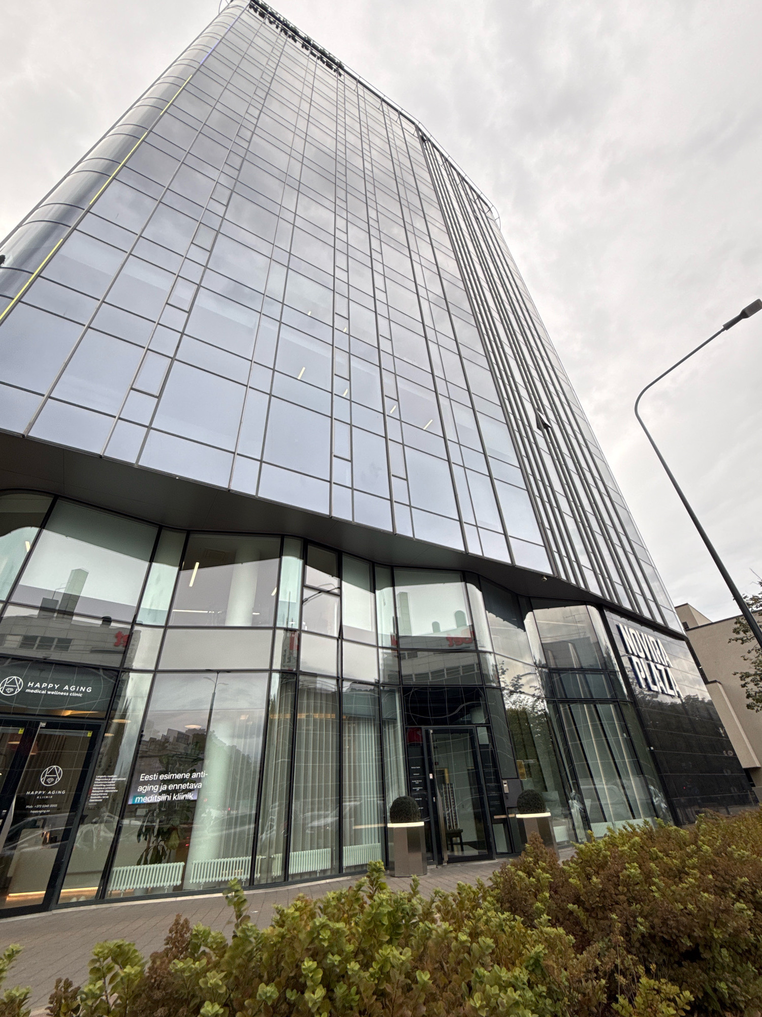 The image shows a modern, glass-fronted office building, identified by signage as "NOVIIRA PLAZA" and "HAPPY AGING medical wellness clinic". The building is located in Tallinn, Estonia. The perspective is from a low angle looking up at the skyscraper, with the sky overcast. In the foreground, there is a paved walkway and dense, low-lying green and brown foliage. A street lamp is visible on the right side of the frame. The entrance to the clinic is visible on the ground floor, featuring glass doors and window displays with informational text. The overall impression is one of an urban, contemporary commercial district.