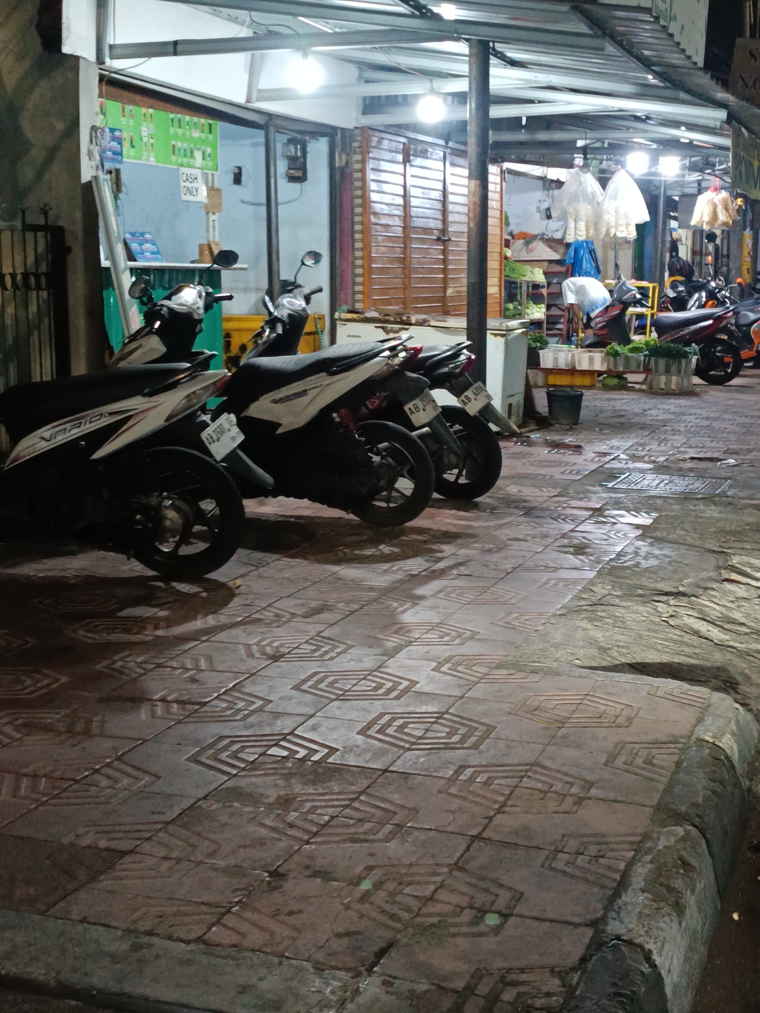 The image depicts a street-level view of a store or market stall in Yogyakarta, Indonesia, at night. The scene is characterized by the presence of several parked motorcycles, indicative of a common mode of transportation in the region. The ground is paved with patterned tiles, some of which appear wet, suggesting recent rain or cleaning.
In the background, the store features a counter with a sign that says "CASH ONLY". Above the counter, a green board displays a grid of images, possibly of products for sale, like beverages or food items. A metal gate is visible on the left side of the image. Further back, various items are displayed on shelves and tables, including what appear to be produce like vegetables, suggesting a food-related business.
The lighting is a mix of artificial lights from the store and ambient street lighting, creating a moderately illuminated but somewhat dark atmosphere. The overall impression is that of a typical commercial street scene in Yogyakarta during evening hours.