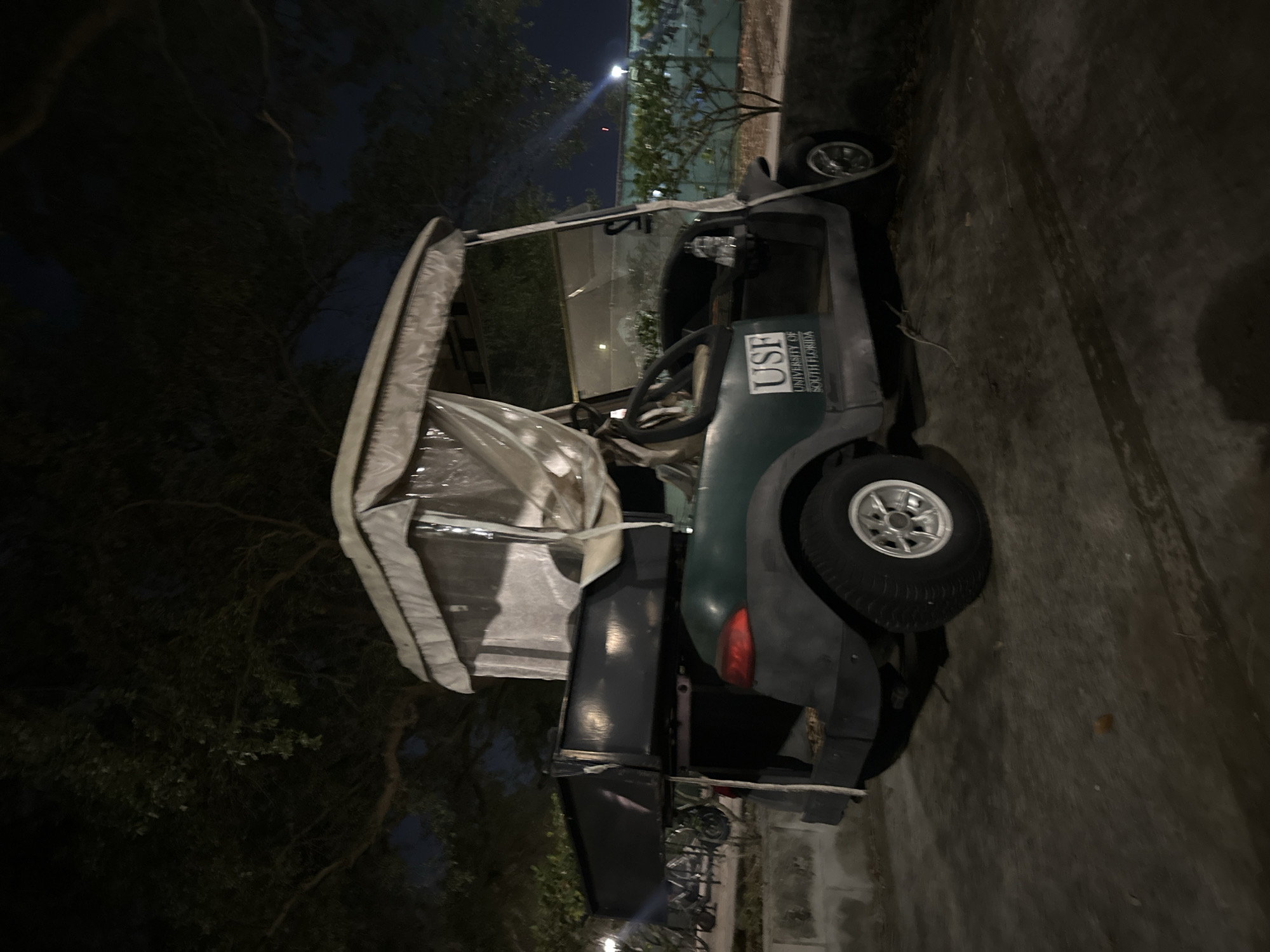 The image depicts a dark green golf cart with a white vinyl roof parked on a paved surface, likely a street or driveway, at night. The golf cart has "USF UNIVERSITY OF SOUTH FLORIDA" printed on its side. Trees are visible in the background. Given the text on the side of the golf cart, the location is likely the city of Tampa, Florida, United States of America where the University of South Florida is located. There are no people visible in the image. The golf cart is the main subject of the image, and it appears to be stationary. The environment is dark, with some ambient light illuminating the golf cart and surrounding vegetation.
