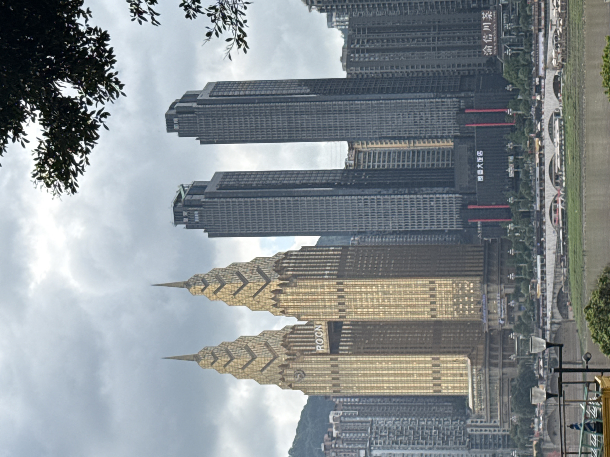 The image displays a dense urban cityscape, likely Yuzhong District, China, under an overcast sky during the daytime. In the mid-ground, two prominent golden-hued, art deco-style skyscrapers with distinctive pointed spires stand out. The text "ROCN" is visible on one of these golden buildings. Behind them, two modern, tall grey rectangular skyscrapers with grid-like facades are visible; one displays the Chinese characters "西南大学" (Southwest University). Further back, another building shows "国大酒店" (Grand Hotel), and to the right, "渝信川菜" (Yuxin Sichuan Cuisine) is visible on a building.
A river or wide waterway runs along the right side of the image, bordered by a long bridge or embankment with decorative arches and streetlights. Numerous other grey residential or commercial buildings extend into the background, creating a layered urban landscape. Lush green foliage from a tree frames the upper left corner of the scene. In the bottom right foreground, a dark metal structure with two white lamp shades is partially visible. No people or specific activities are discernible, presenting a static view of the city's architecture and natural setting.
