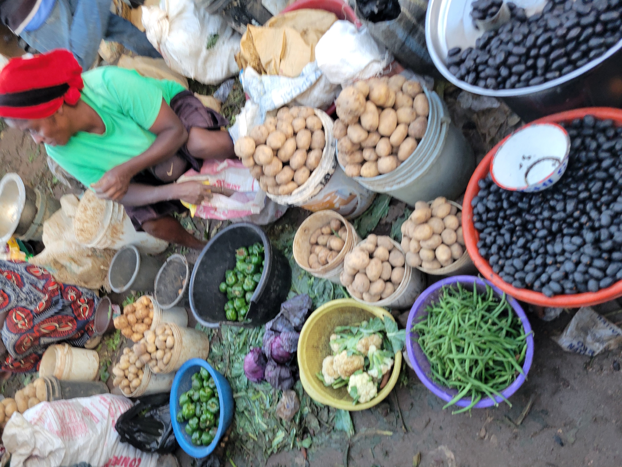 The image depicts an outdoor market scene, likely in Gura Top, Nigeria. A woman wearing a green t-shirt and a red headscarf is seated, engaged in selling produce. In front of her are various vegetables arranged in buckets and bowls. To the left, a blue bowl contains green bell peppers, and a larger black bucket also holds green bell peppers. A yellow bowl displays cauliflower florets, and to the left of that, several heads of red cabbage are visible. Behind the cabbage, piles of potatoes are stored in white buckets. In the foreground, a purple bowl is filled with green beans, and to its right, an orange bowl overflows with dark, small berries, possibly dates. Another metallic container to the right also contains a similar dark berry-like fruit. Bags of produce and other market goods are scattered around the scene, with a patterned cloth draped over a person in the background. The ground appears to be dirt or mud, typical of an open-air market.