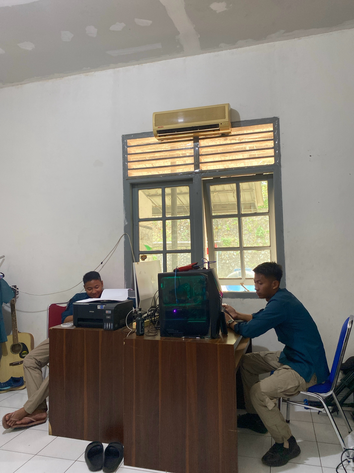 The image depicts an indoor scene, likely an office or workspace, in Tanjung Pinang, Indonesia. Two young men are present. The man on the left is seated at a wooden desk, wearing a dark blue shirt and khaki pants, and appears to be working on a printer. His feet are bare, clad in flip-flops. An acoustic guitar is leaning against the wall to his left, and the desk has a black EPSON printer on it. The man on the right is also seated at a similar desk, wearing a dark blue shirt and khaki pants. He is actively engaged with a computer, with his hands on the keyboard and screen. A gaming-style computer tower with illuminated internal components is visible on his desk, along with various cables and accessories. A blue-seated chair is positioned at his desk. In the background, a large window with horizontal blinds is visible, and an air conditioning unit is mounted above it. The walls are painted white, and the ceiling shows signs of repair work with plaster patches. The floor is tiled. The overall impression is a functional, if somewhat unfinished, workspace.