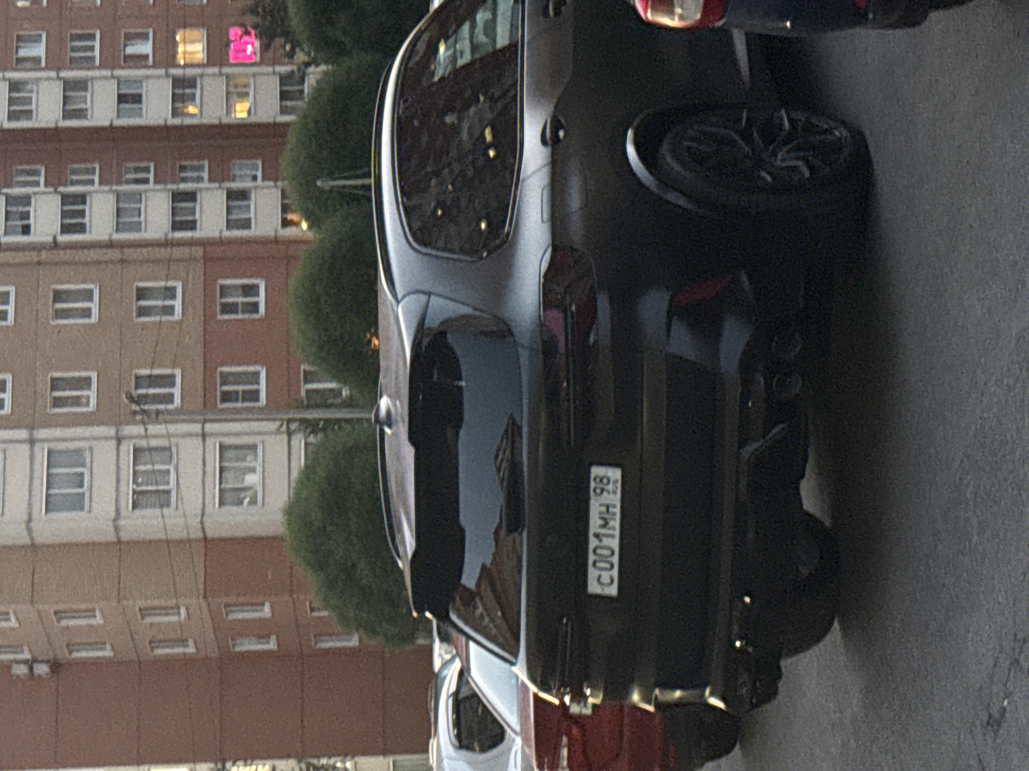 An outdoor, eye-level, slightly low-angle, full shot shows the rear passenger side of a black SUV parked on an asphalt surface. The SUV's license plate, partially obscured by glare, reads "C001MH 98". The SUV is dark gray or matte black with tinted windows and black rims. The building behind the SUV is a multi-story residential building, brick on the left side with white and beige sections on the right. There are trees visible behind the SUV. The sky is not visible. The image appears to be taken in Saint Petersburg, Russia, as indicated by the license plate format. There are no people visible in the image.