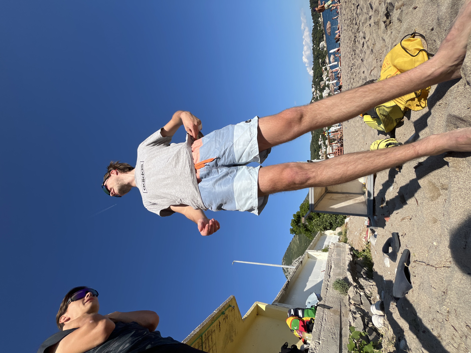 Two young men are on a sandy beach in Zankovići, Montenegro. The man on the left, partially visible and wearing sunglasses, looks down. The man on the right, in focus and also wearing sunglasses, looks up at the sky. He is wearing a grey t-shirt with "adidas" printed on it and light blue and light grey swim shorts. He is adjusting his shorts. A bright yellow bag lies on the sand next to his feet. The background shows a clear blue sky, a building with a yellow awning, a lamp post, vegetation, and the coastline with buildings and people.