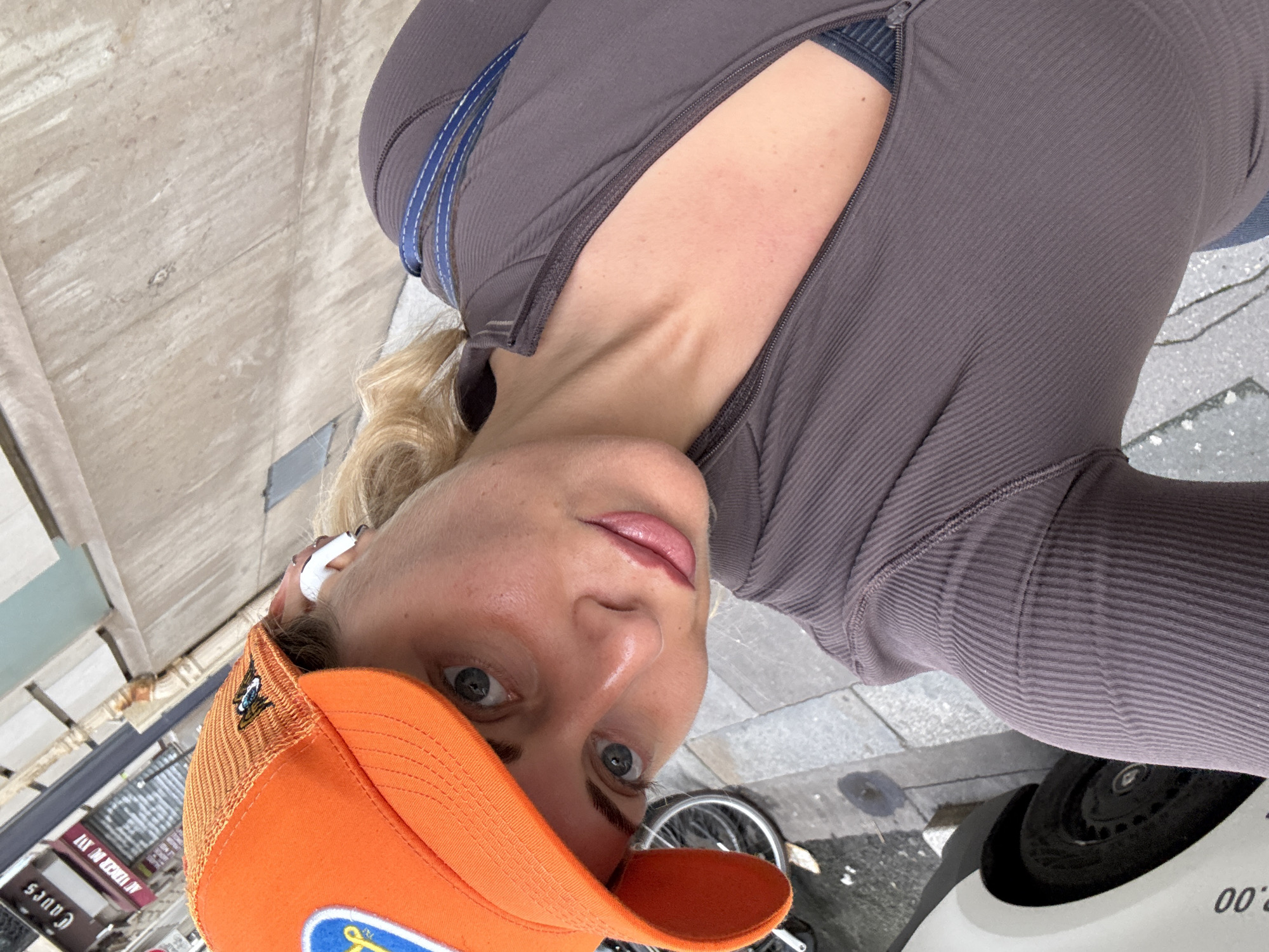 A person wearing an orange baseball cap, a gray ribbed long-sleeve shirt, and an earpiece stands in a city, presumably Paris, France, based on the style of street signs visible in the background. The person appears to be holding the camera at an upward angle, capturing a selfie. A bicycle wheel and the side of a white vehicle are visible in the foreground. The sidewalk is made of concrete tiles. Buildings with signs, including one with "CAUES" written on it, are visible.