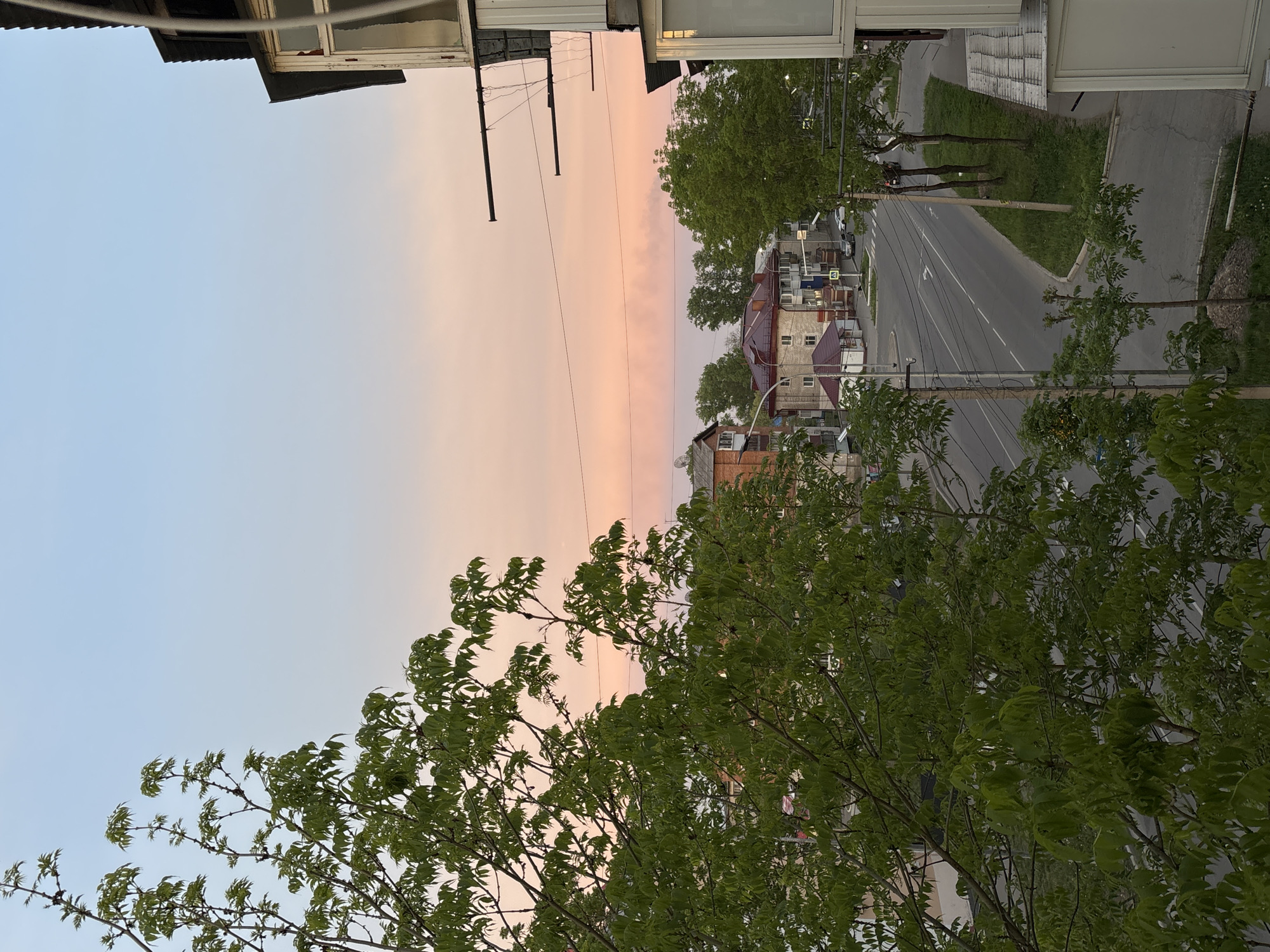 The scene depicts a view from an elevated position, likely a window or balcony, overlooking a residential street in an urban area of Unknown, Unknown. The sky is visible, transitioning from a light blue to a pinkish hue, suggesting either sunrise or sunset. Power lines run along the street, connecting to utility poles. Buildings line the street, varying in height and architectural style, with some appearing to be two or three stories tall. A paved road runs between the buildings, marked with white lines indicating traffic lanes. Green trees are present, both along the street and in the foreground, with leaves in full bloom. The foreground also features a section of a building, presumably where the image was taken from, with some architectural details visible.