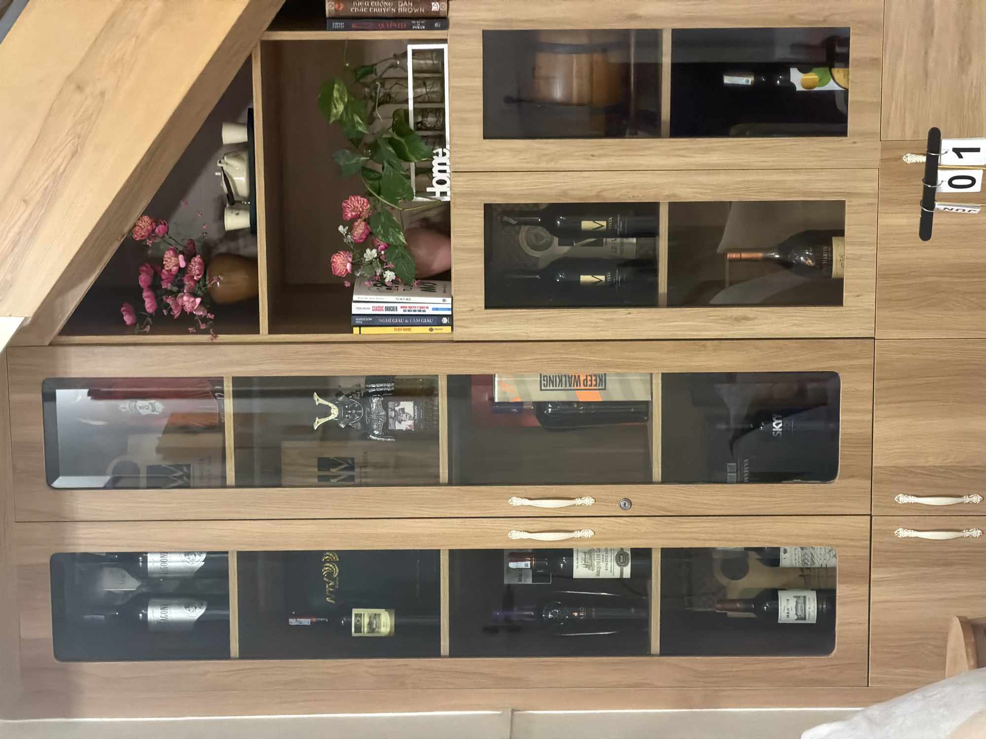 The image displays a multi-functional wooden display cabinet, likely situated in a home in Ninh Chu Ward, Vietnam. The cabinet, made of light-colored wood, features a combination of open shelves and glass-fronted compartments, primarily used for storing beverages and books, along with decorative items.
On the far left, a triangular section, possibly under a sloped ceiling or staircase, houses a decorative ceramic teapot set and a vase filled with pink artificial flowers. Adjacent to this, an open shelf holds a stack of books, including titles in Vietnamese such as "ĐẮC NHÂN TÂM" (How to Win Friends and Influence People), "NGHỆ THUẬT QUYẾN RŨ" (Art of Seduction), along with a green potted plant and a white decorative frame spelling "Home".
The upper and middle sections of the cabinet feature numerous glass-fronted compartments filled with a variety of wine bottles. Visible labels include "DRAGONI," "MARTIN," "MARQUISE EDE LAI," and "VISTA." A bottle of "SKYY" vodka and a box labeled "KEEP WALKING" are also present. A small wooden barrel is visible in one of the top compartments. Below the glass-fronted sections are several drawers with ornate white handles, one featuring a keyhole. More books are stacked on an open shelf, including "CLASSIC DRUCKER" and Vietnamese titles like "TƯ DUY MỚI" (New Thinking), "NGHĨ GIÀU & LÀM GIÀU" (Think and Grow Rich), and "TƯ DUY NGƯỢC."
The bottom section continues with glass-fronted shelves displaying more wine bottles. On the far right vertical panel of the cabinet, two more drawers with similar handles are visible, and a hanging date display shows "01" and "JUN" (June), indicating the first day of June. The scene depicts a static, organized display of personal belongings.