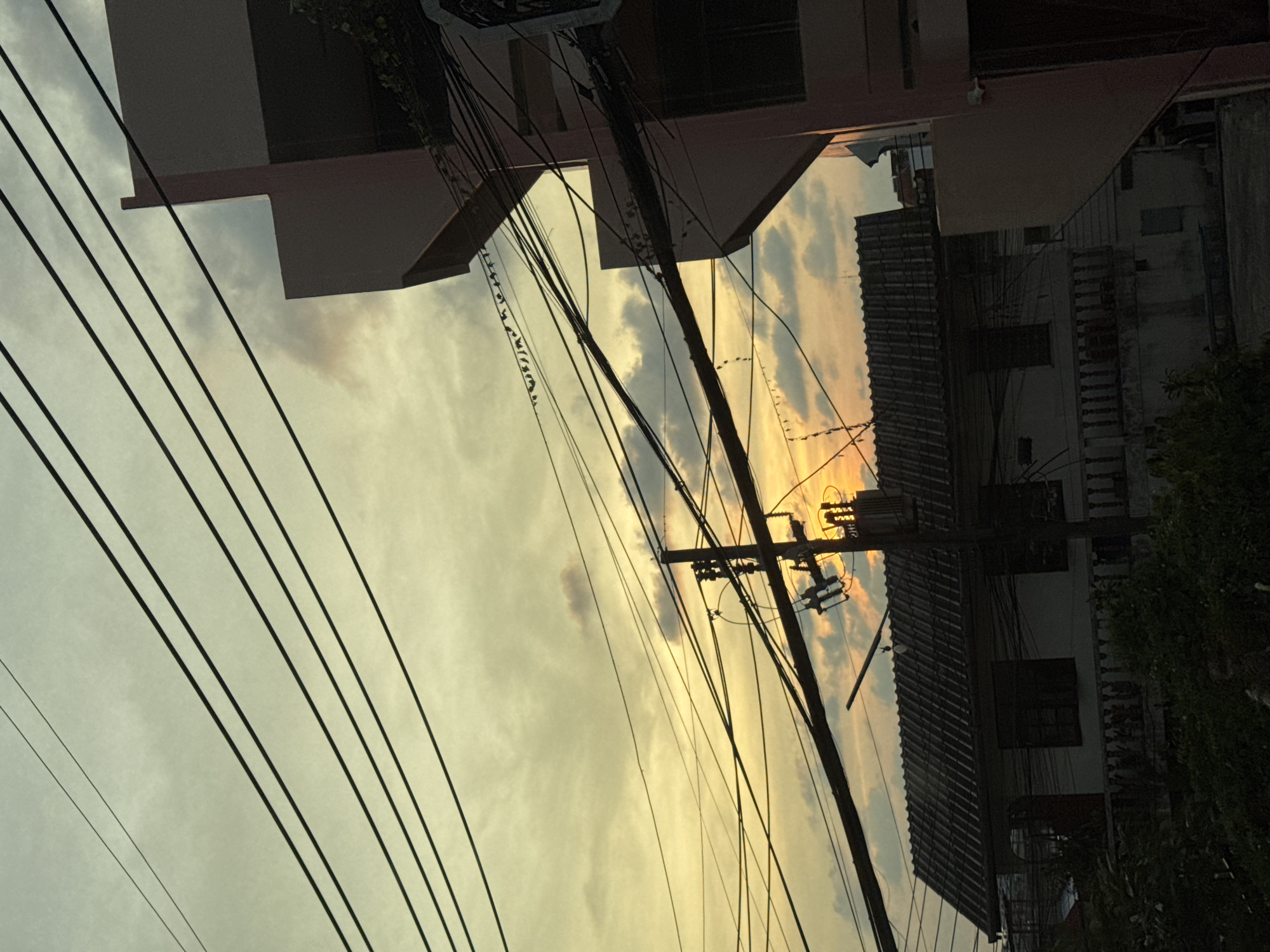 This eye-level shot is of Patong, Thailand. It focuses on the sky at sunset. In the foreground are several electrical wires extending to the left. In the middle ground is a utility pole. Behind the utility pole is a sunset of oranges and yellows. Buildings surround the sky.