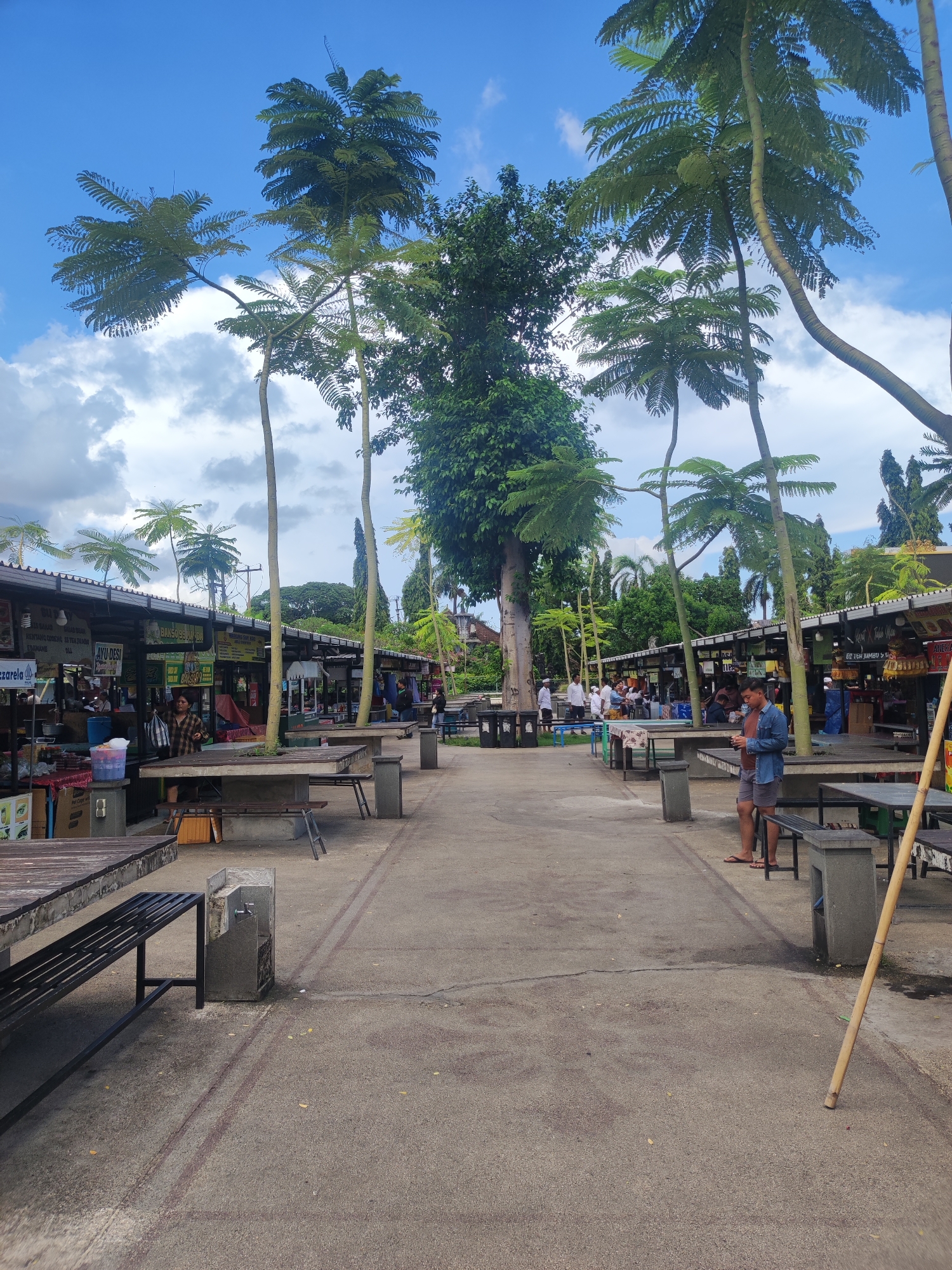 The image depicts a marketplace in the city of Gianyar, Indonesia. The marketplace is open-air and features numerous stalls along either side of a wide, paved pathway. Many people are present. Some are at the stalls, while others are sitting at tables. The tables and benches are concrete with wooden tops and seats. The stalls have dark, overhanging roofs. A man is standing on the right-hand side of the pathway. He wears a blue shirt and shorts. He is looking at his phone. Several tall, thin trees with fern-like leaves grow among the stalls and in the central area. A large, leafy tree stands at the center of the image. The sky is blue with scattered clouds.