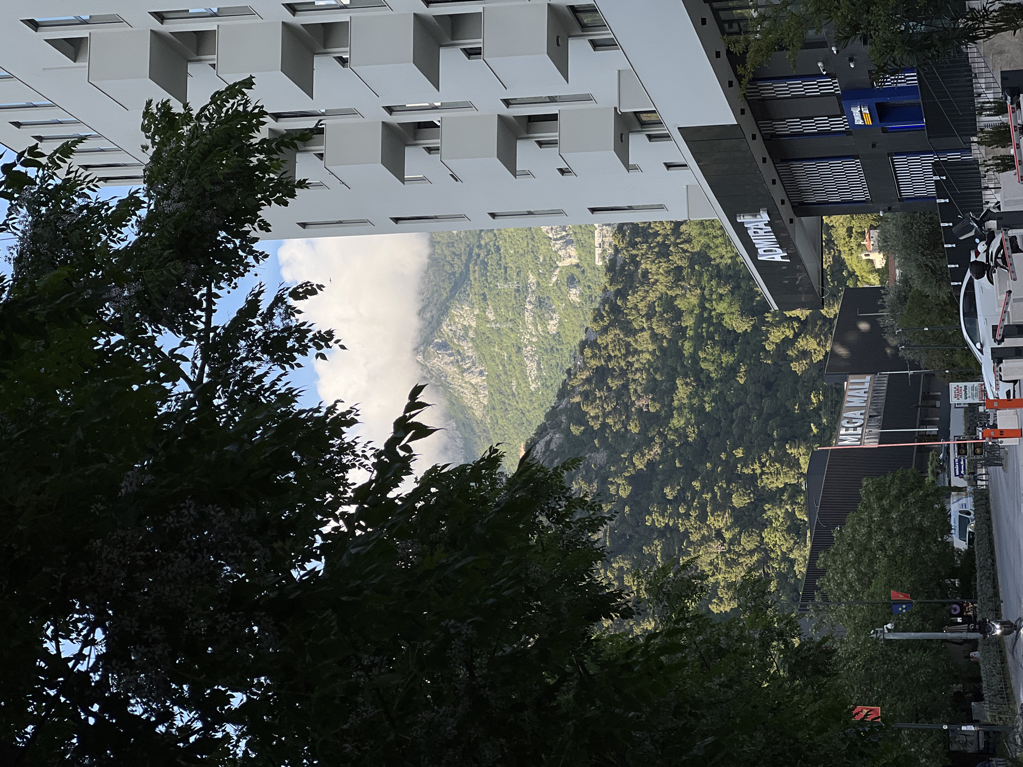 The image shows a view in Budva, Montenegro, looking towards a mountain landscape partially obscured by clouds. In the foreground are trees. Above the trees is a multi-story, white building with a distinctive balcony design. Adjacent to this building is a darker structure with "ADMIRAL" emblazoned on its side. Further in the distance, the MEGA MALL can be seen. The street is visible with cars and street lights.